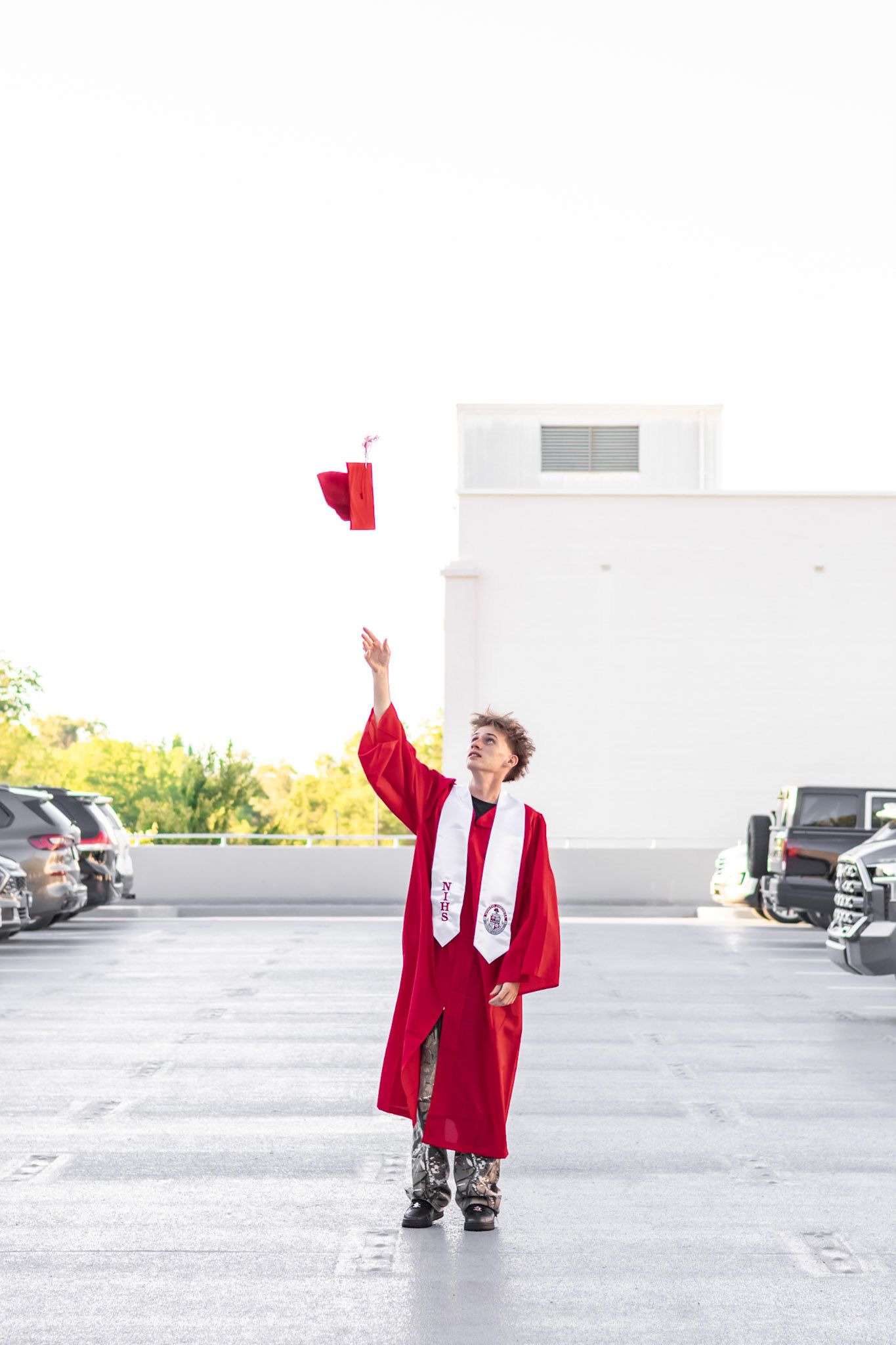 A young male graduate in a red cap and gown tosses his cap into the air on a rooftop parking lot.