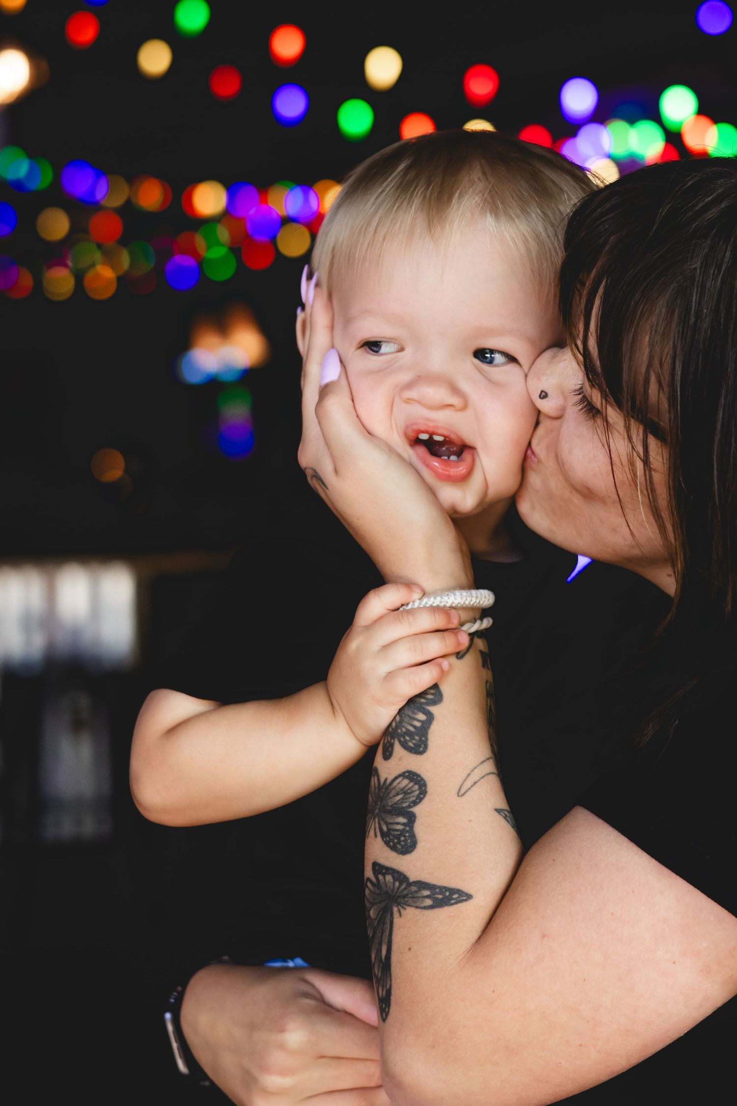 Mother kissing smiling baby boy on cheek with colorful bokeh lights glowing in dark background.