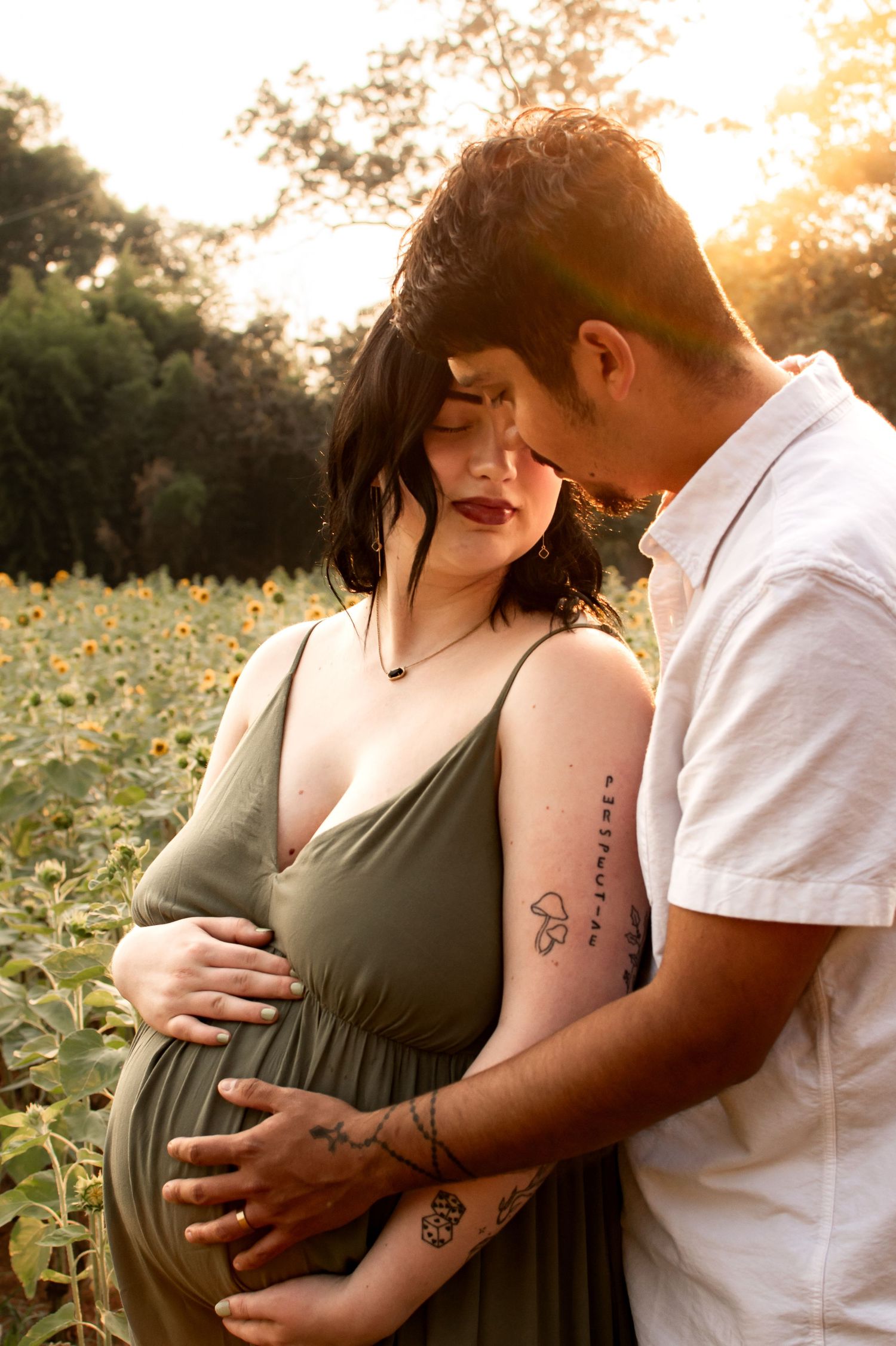 Pregnant couple embracing in sunflower field at golden hour, woman in olive green dress cradling baby bump.