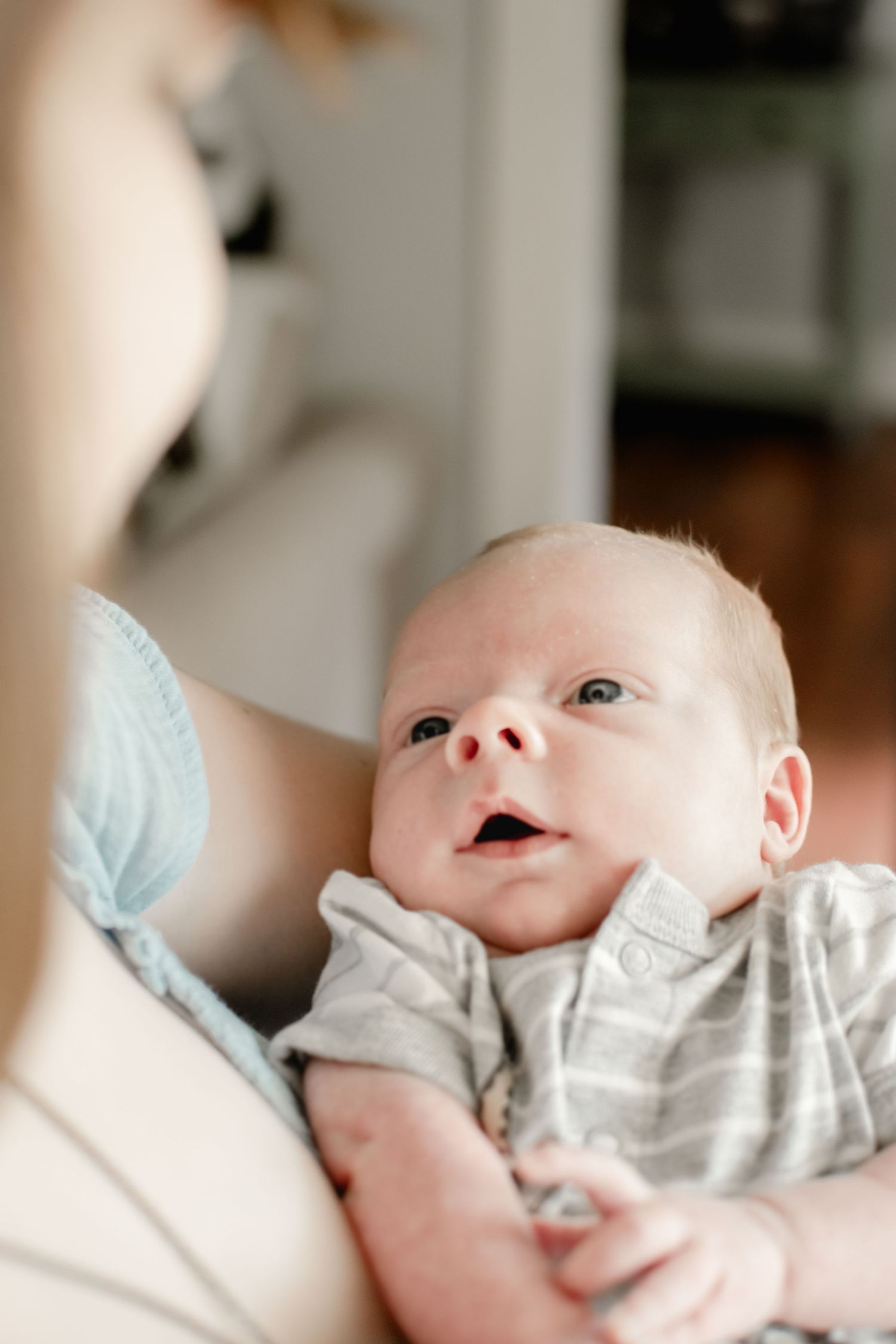 A newborn baby with wide eyes looks up while being cradled in a parent's arms indoors.