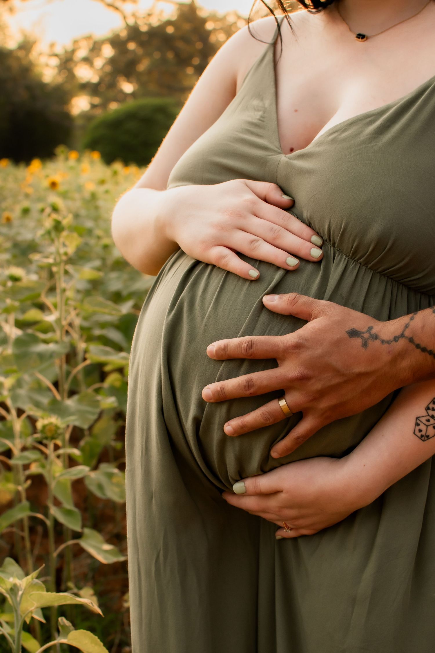 Couple cradling pregnant belly in olive green maternity dress in sunflower field at sunset.