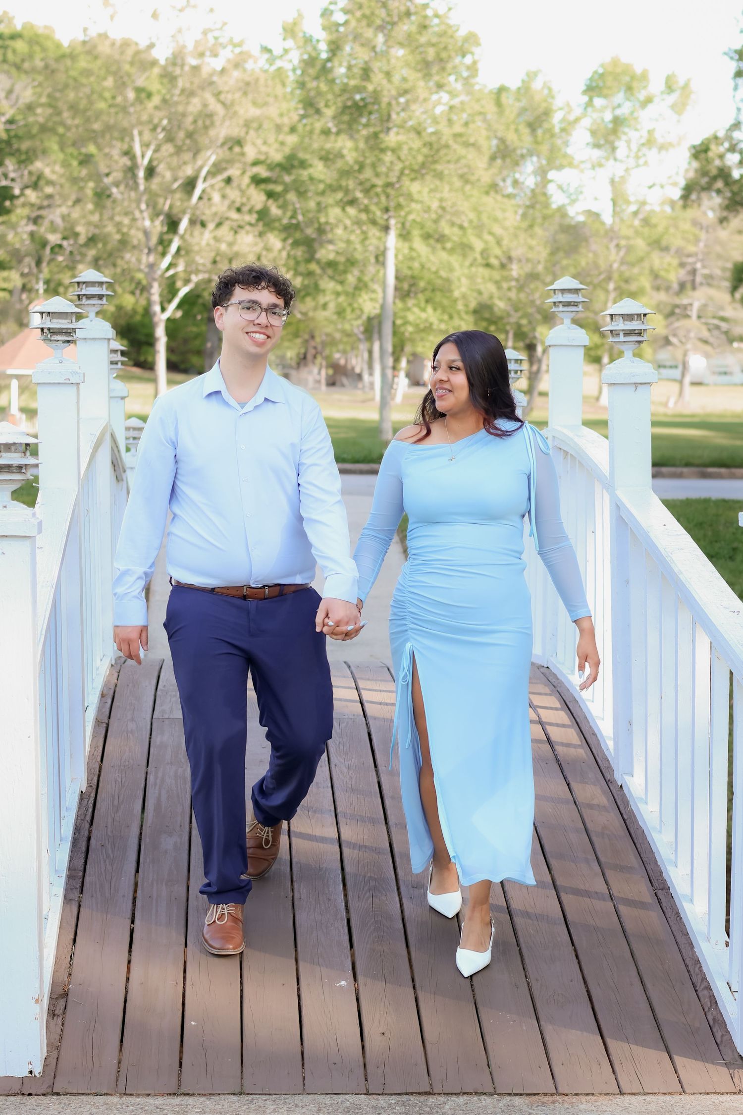 Happy couple in matching light blue outfits holding hands while walking across white wooden bridge.