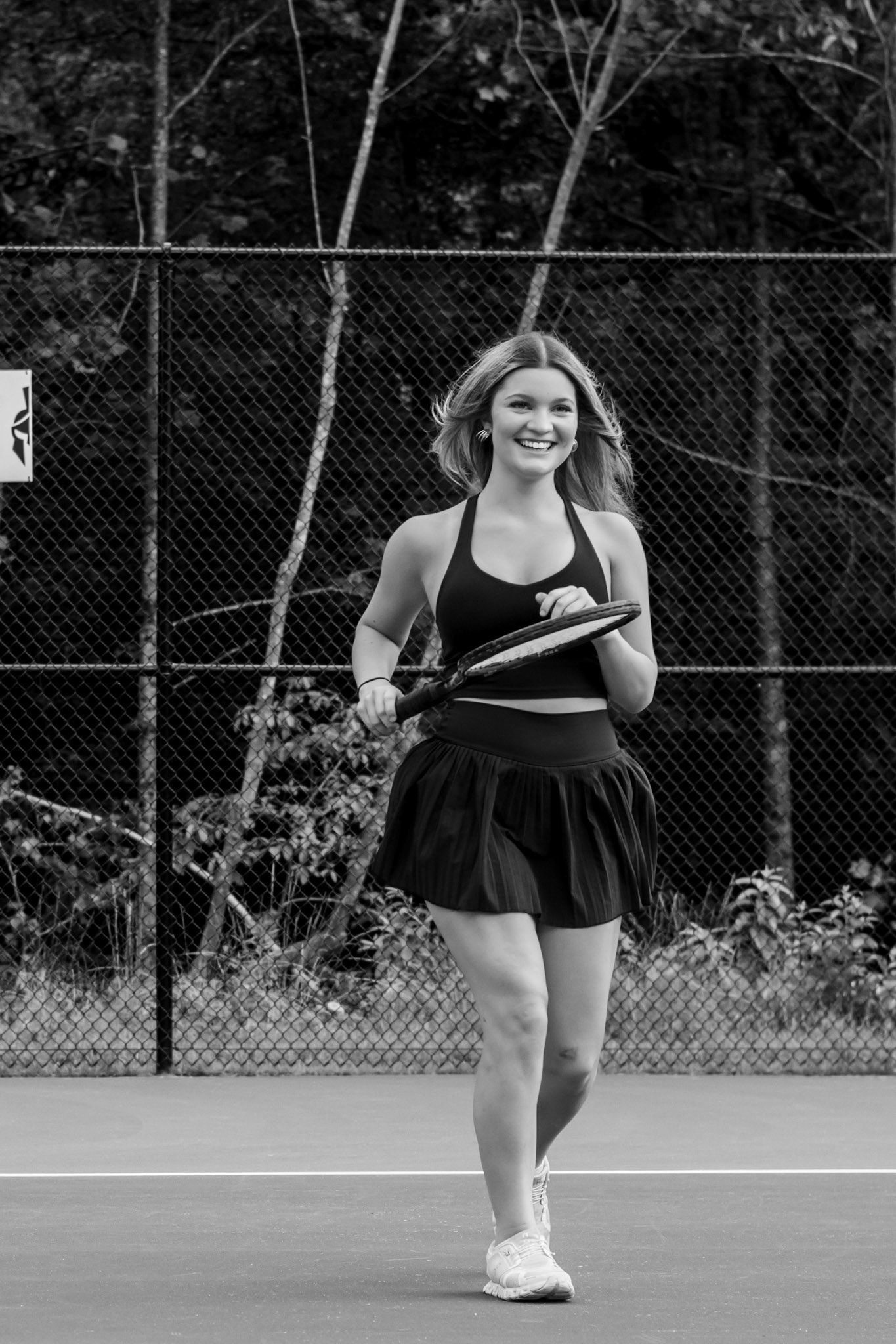 A smiling blonde woman walks on a tennis court in a black skirt and top holding a racket, black and white photo.