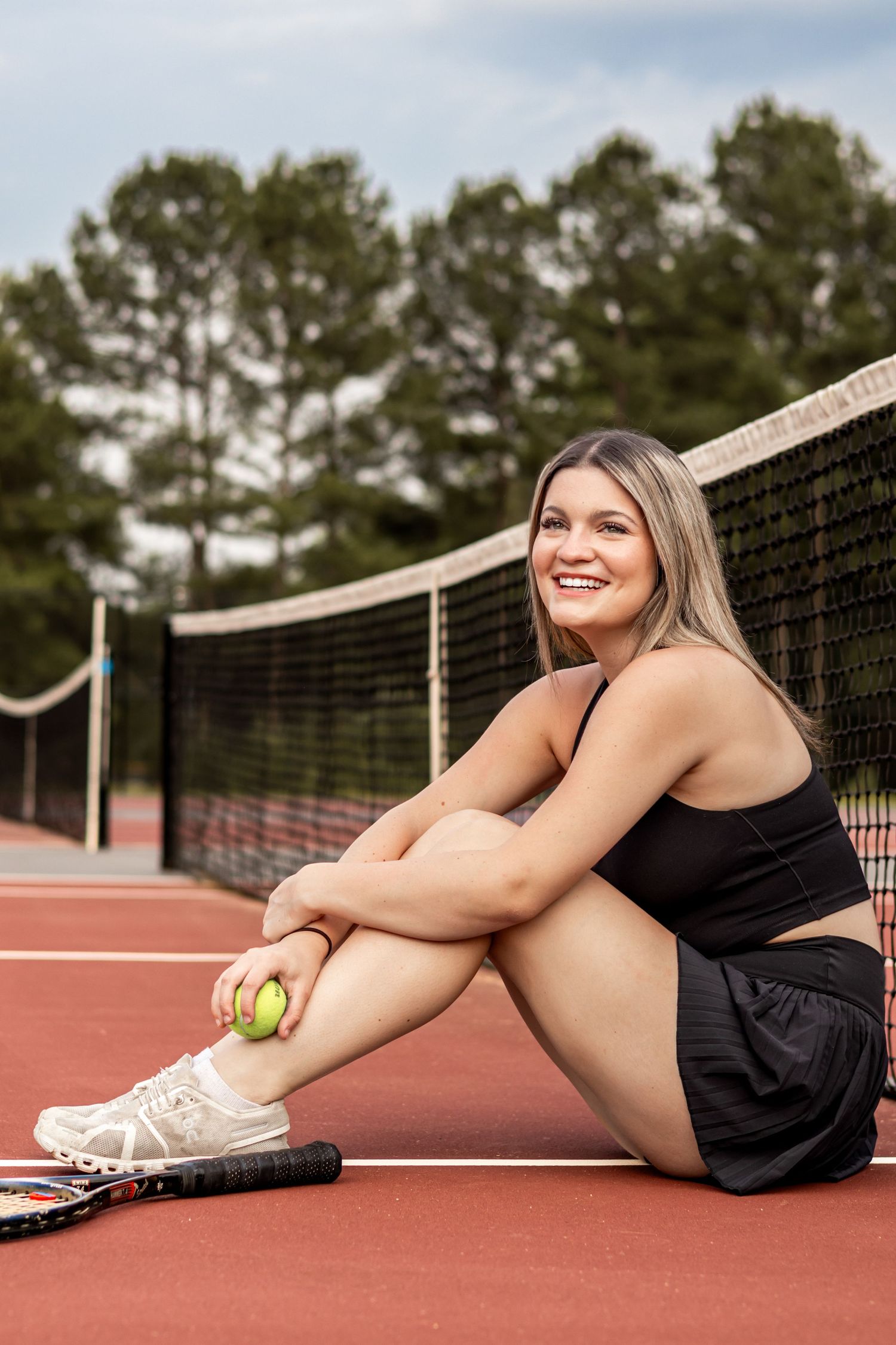 Smiling blonde woman in black tennis outfit sitting on red court holding tennis balls with racket beside her.