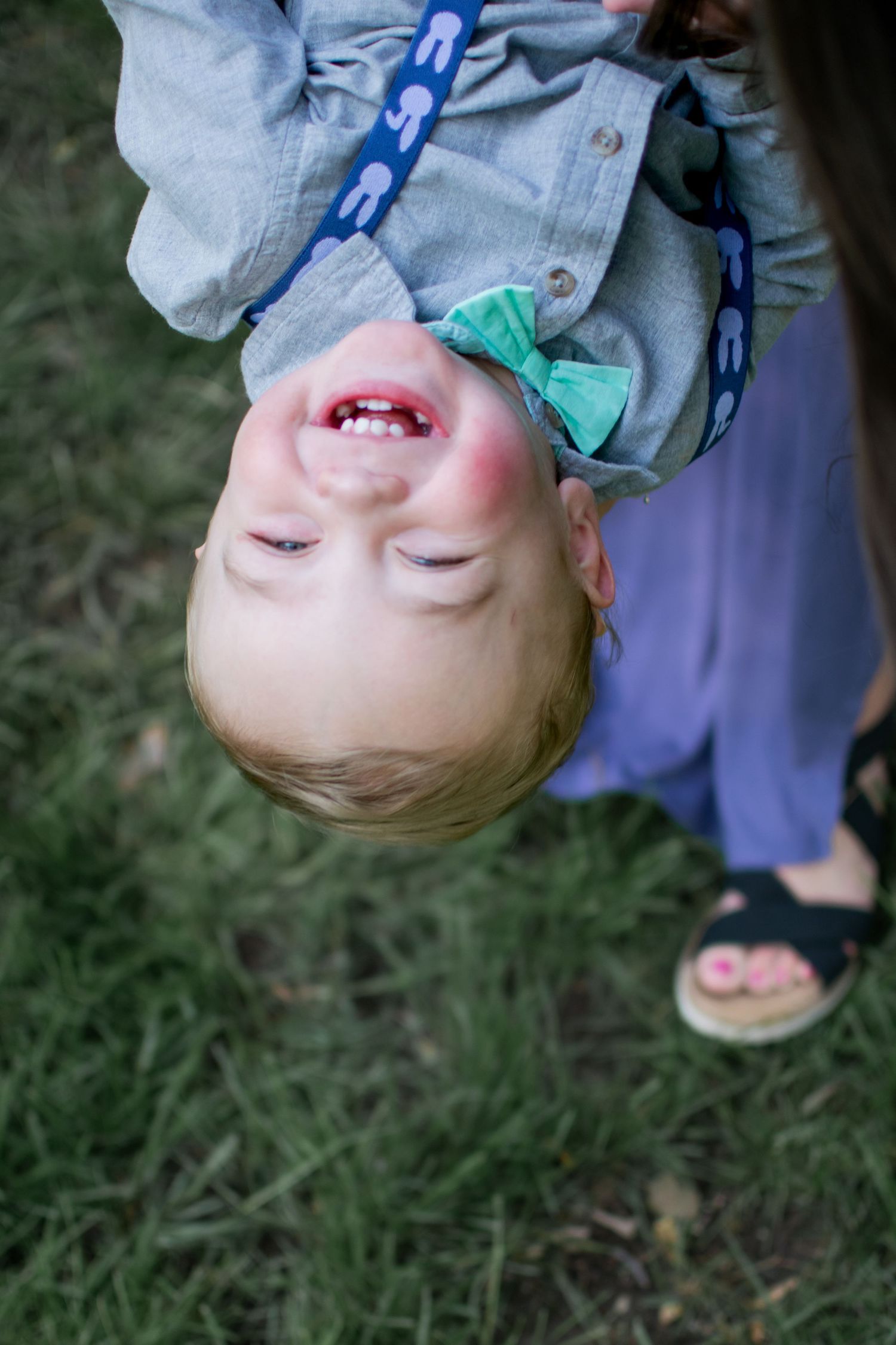 Laughing toddler held upside down wearing denim shirt and green bow headband outdoors.