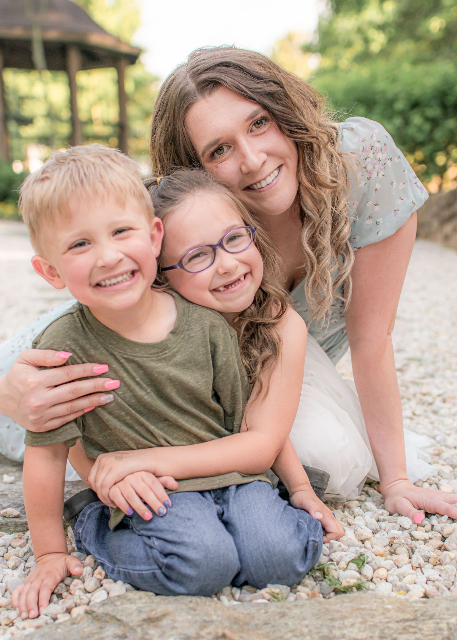 A smiling mother hugs her two young children, a boy and a girl with glasses, on a pebble path outdoors.