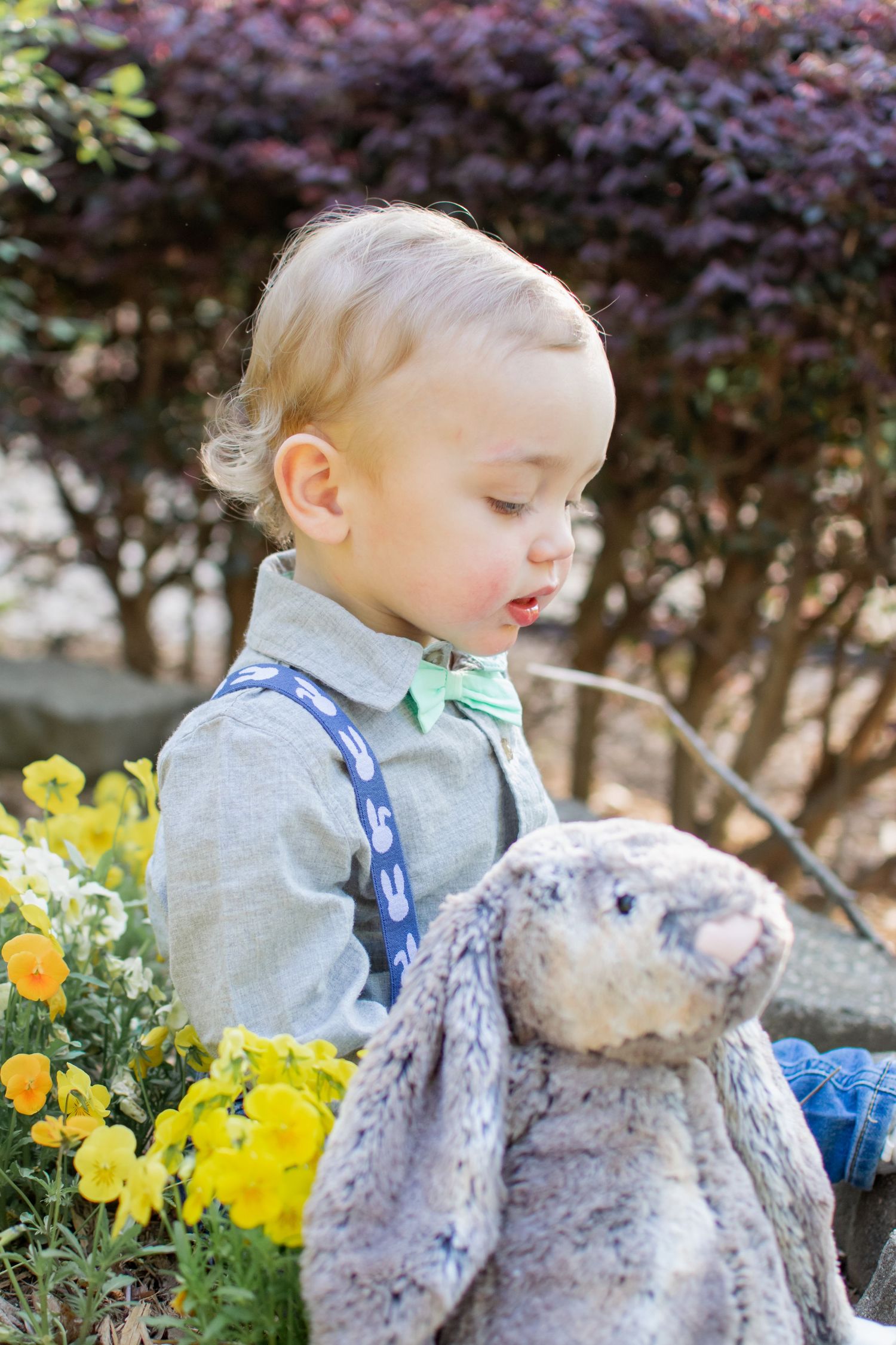 Adorable blonde toddler boy in suspenders and bow tie holding a stuffed bunny among yellow flowers.