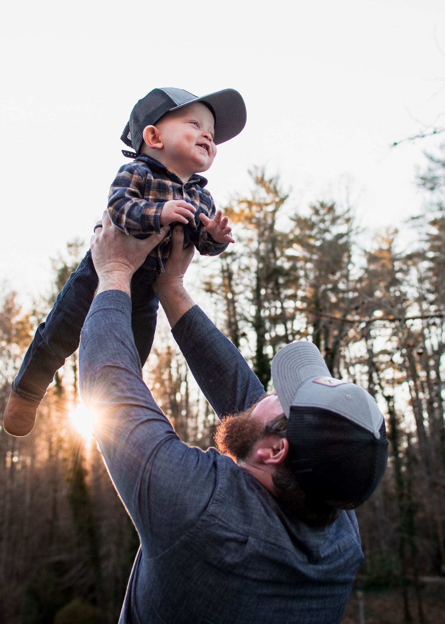 Joyful father lifting laughing baby boy in plaid shirt overhead at sunset in woods.