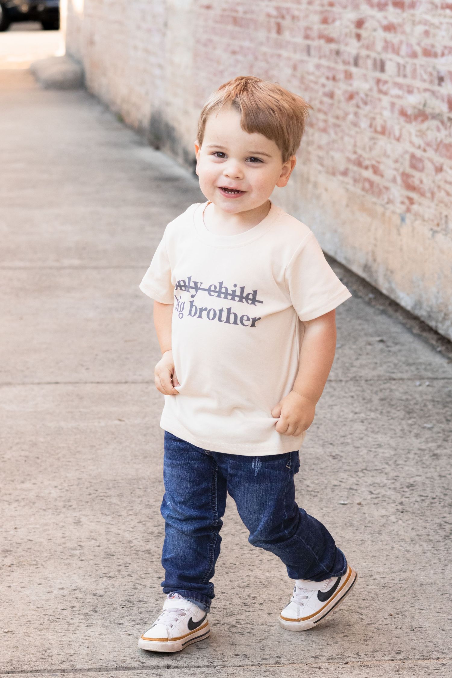 Adorable toddler boy wearing big brother t-shirt and jeans walking on sidewalk near brick wall.