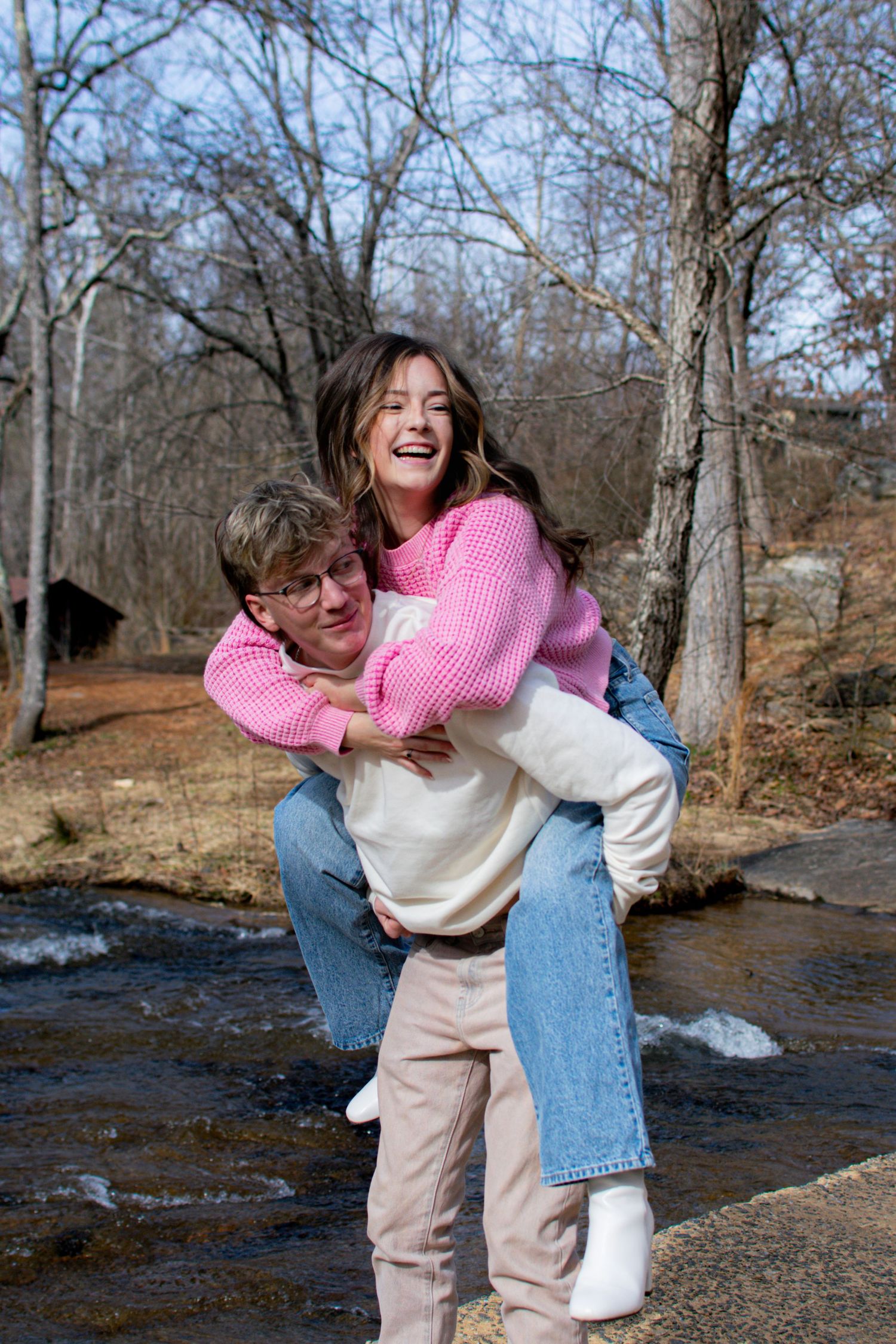 Happy young couple laughing as man gives woman piggyback ride near creek in winter.