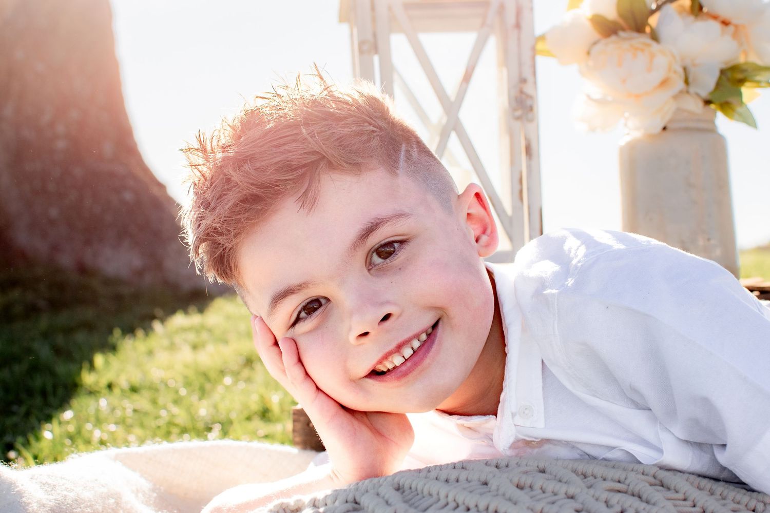 A smiling young red-haired boy resting his chin on his hand while lying on a blanket outdoors in sunlight.