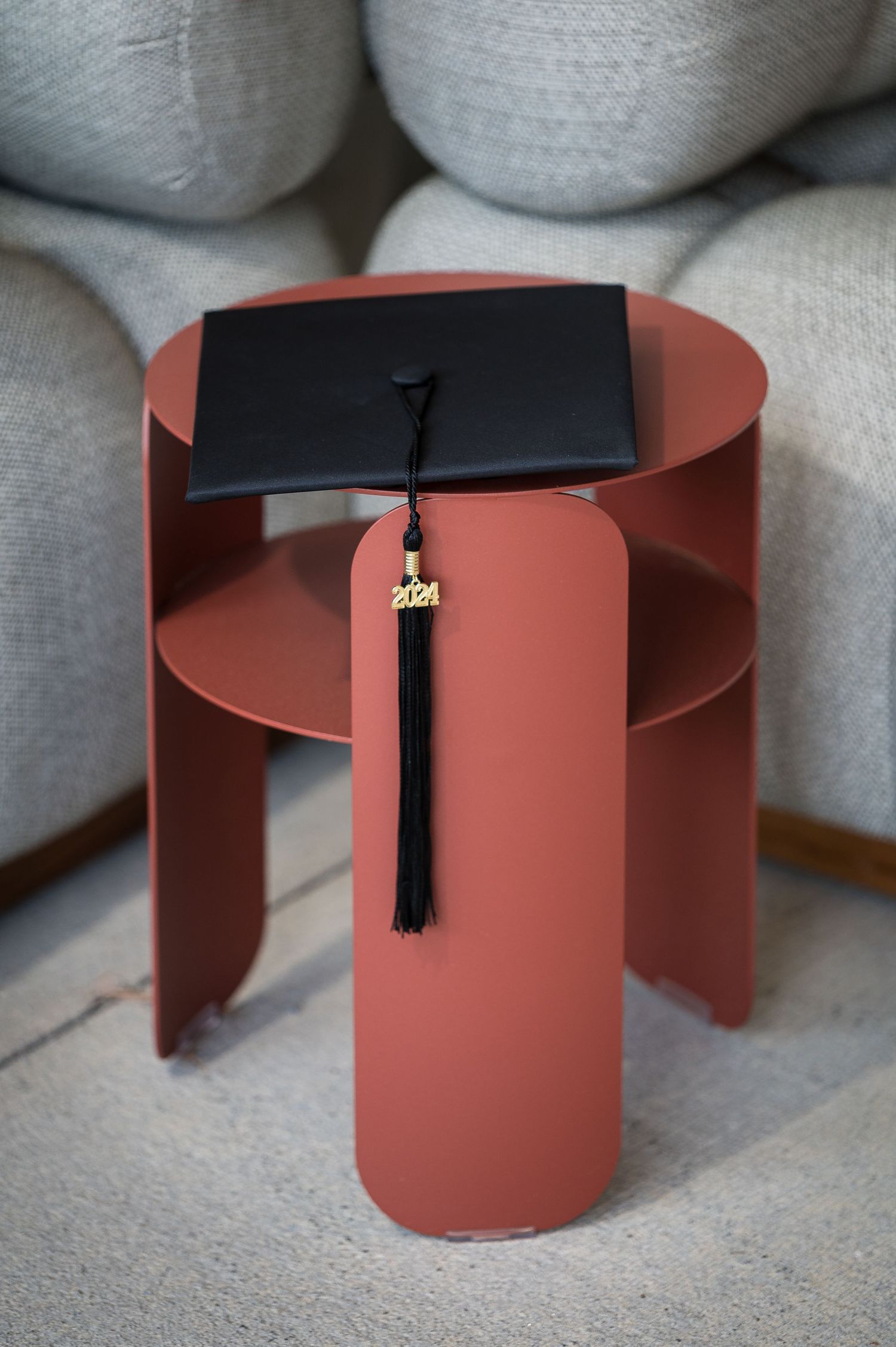 A graduation cap with tassel sits on a modern coral-colored side table against white cushions.