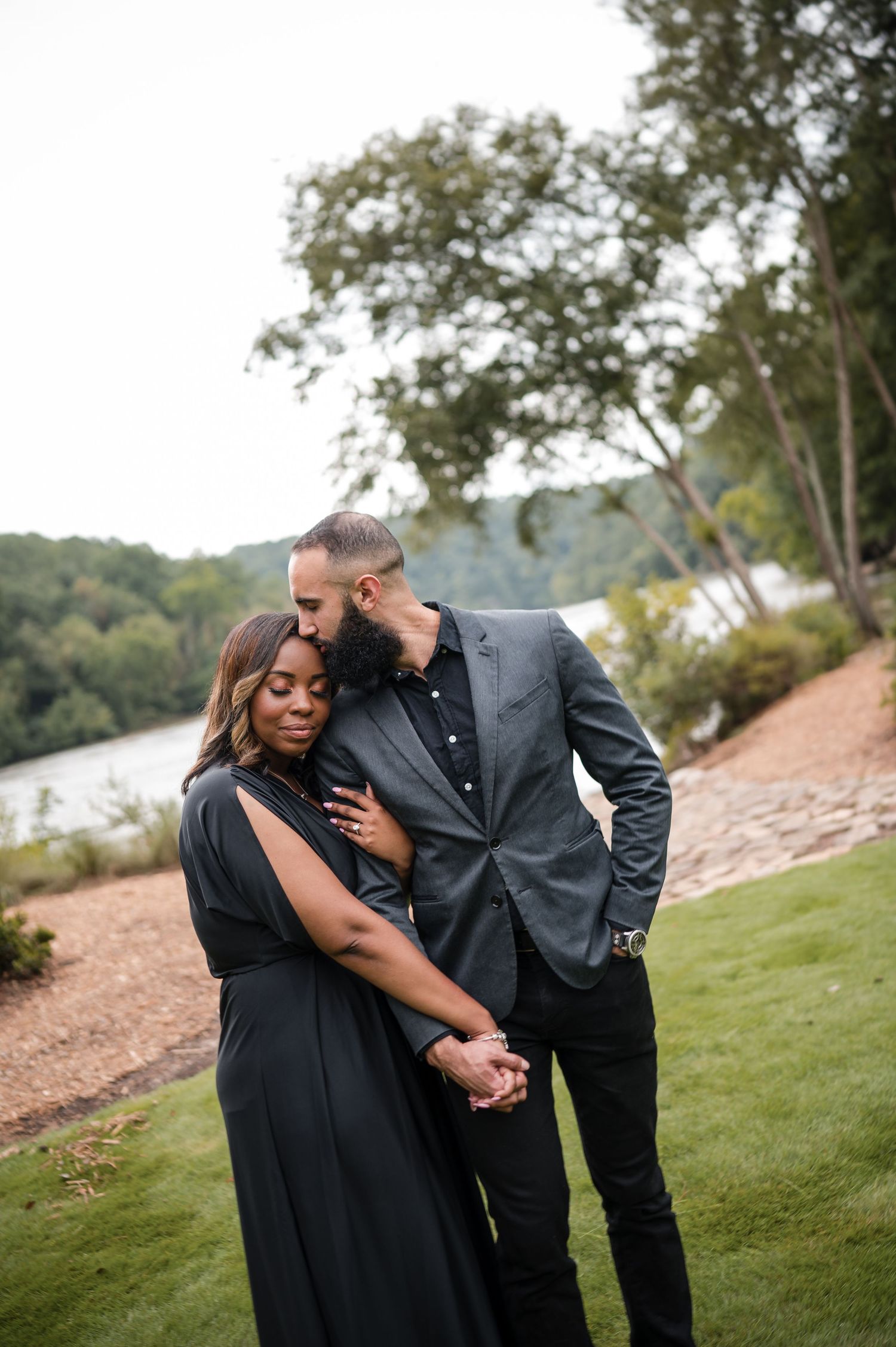 A romantic couple embraces on a grassy hillside overlooking a scenic lake at sunset.