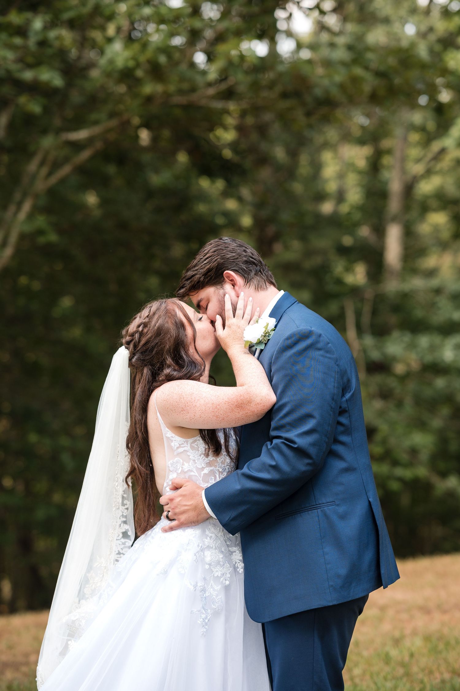 A romantic first kiss scene between newlyweds on a grassy hillside with trees in the background.