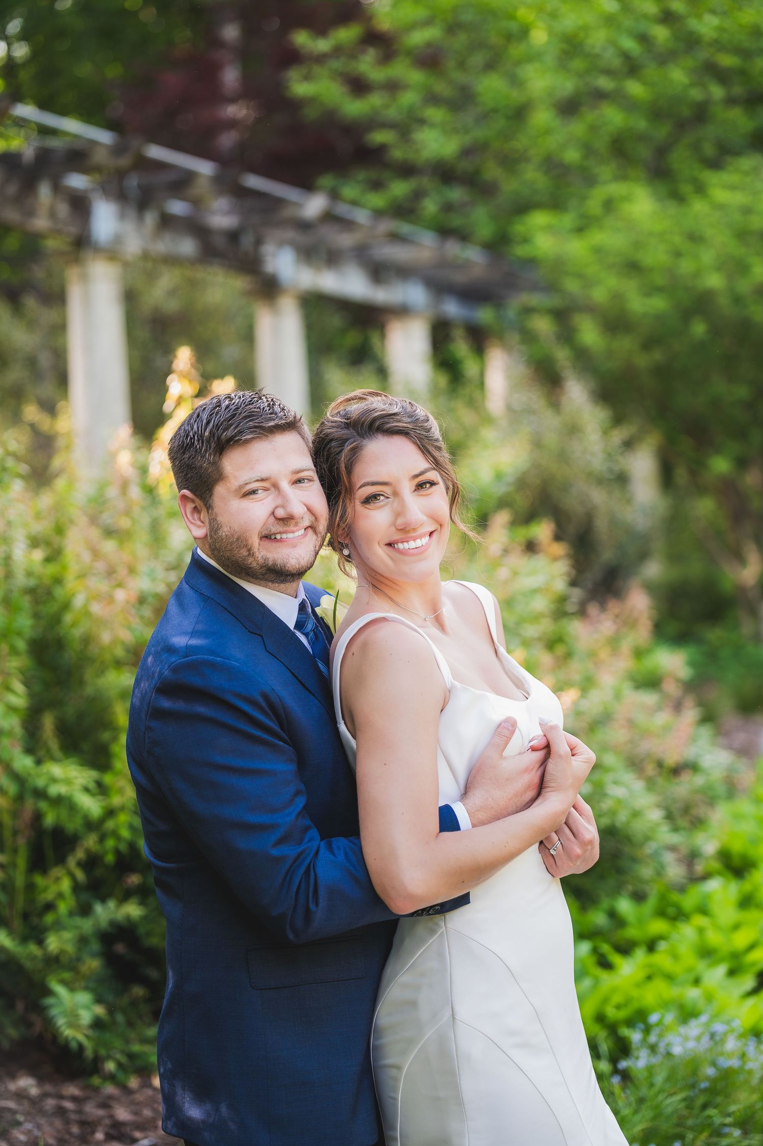Couple in wedding attire embrace and smile in a romantic outdoor garden setting with greenery.