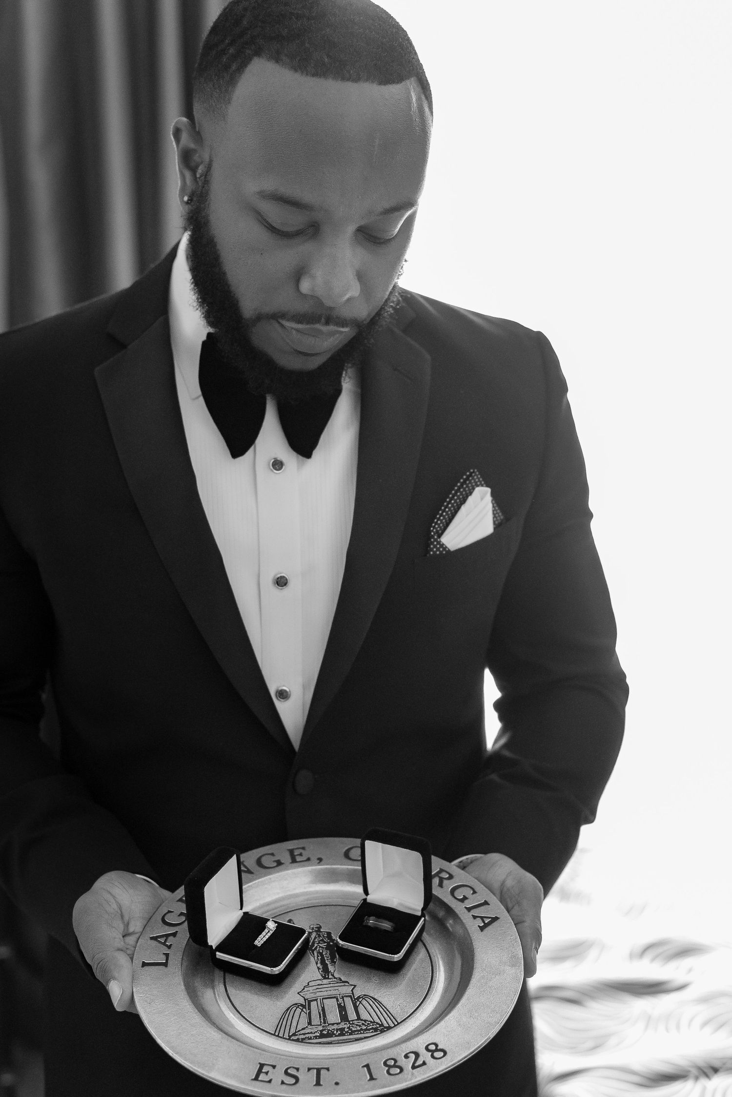 Groom in black tuxedo examining ring box in classic black and white photograph.