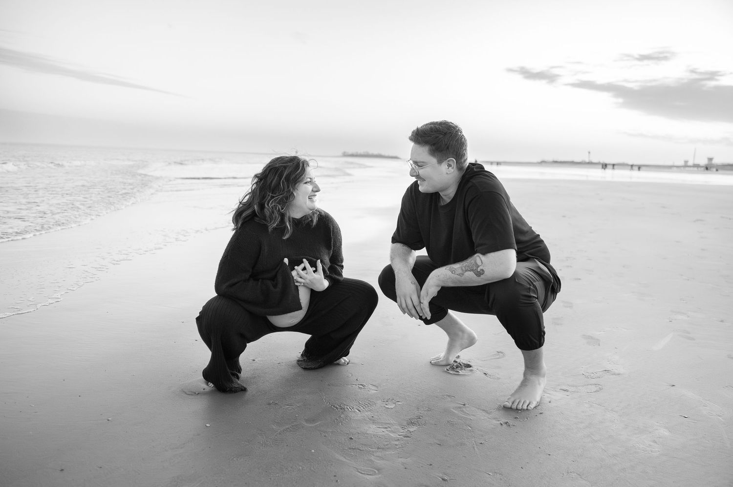 Black and white photo of two people crouching and talking on a beach at sunset.