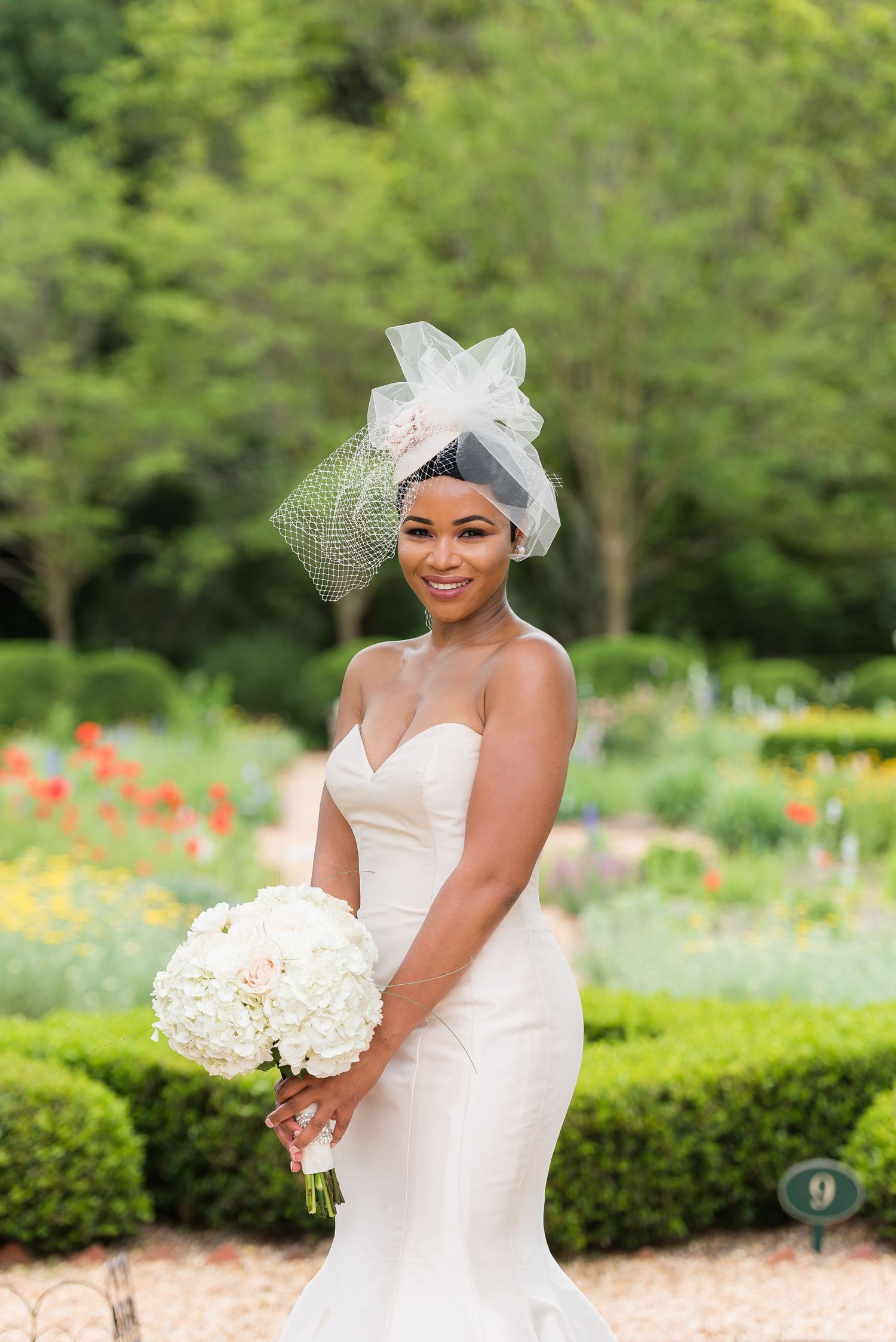 Bride in white dress and veil poses in garden with colorful flowers and manicured hedges.