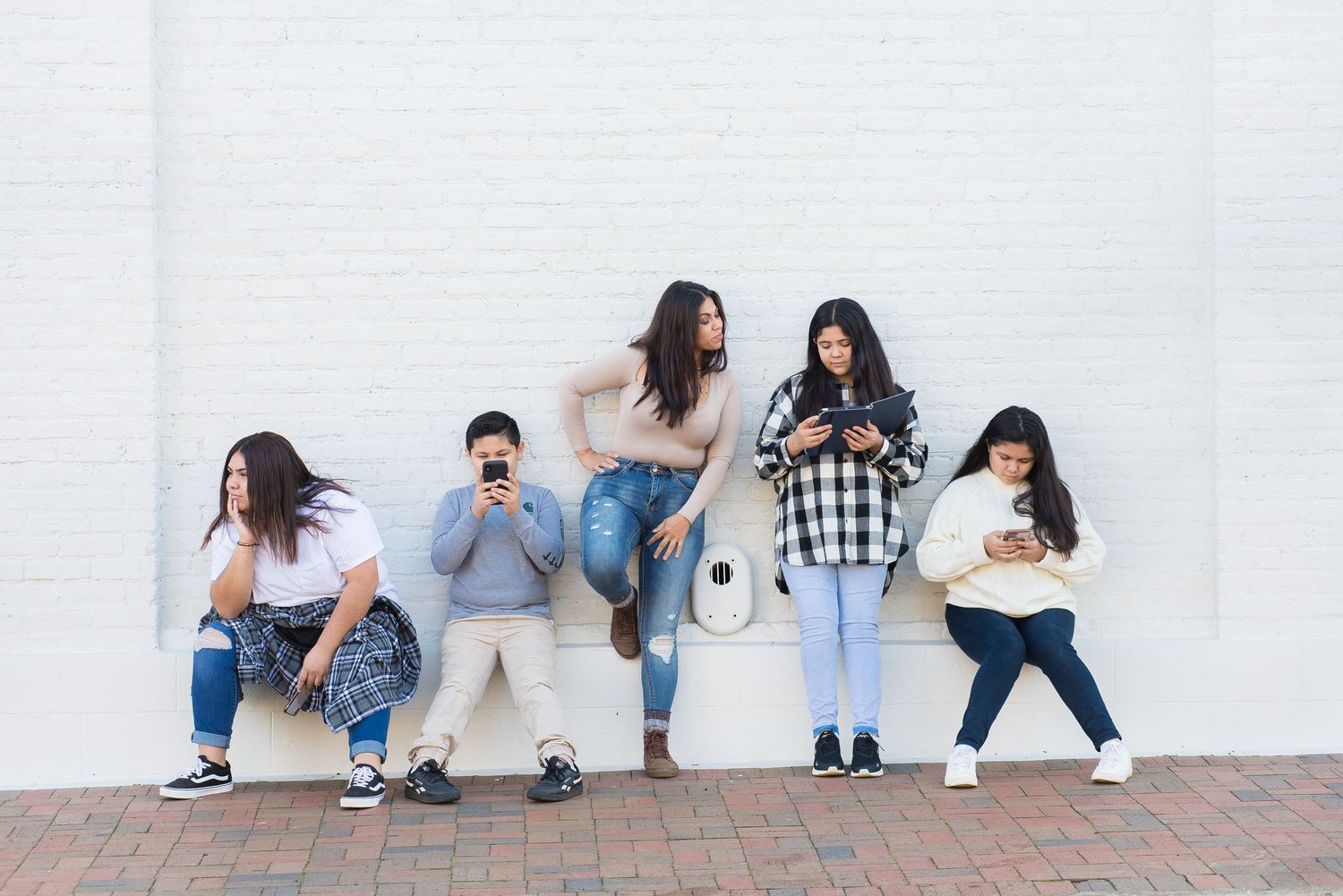 Five people sit against a white wall looking at their mobile phones.