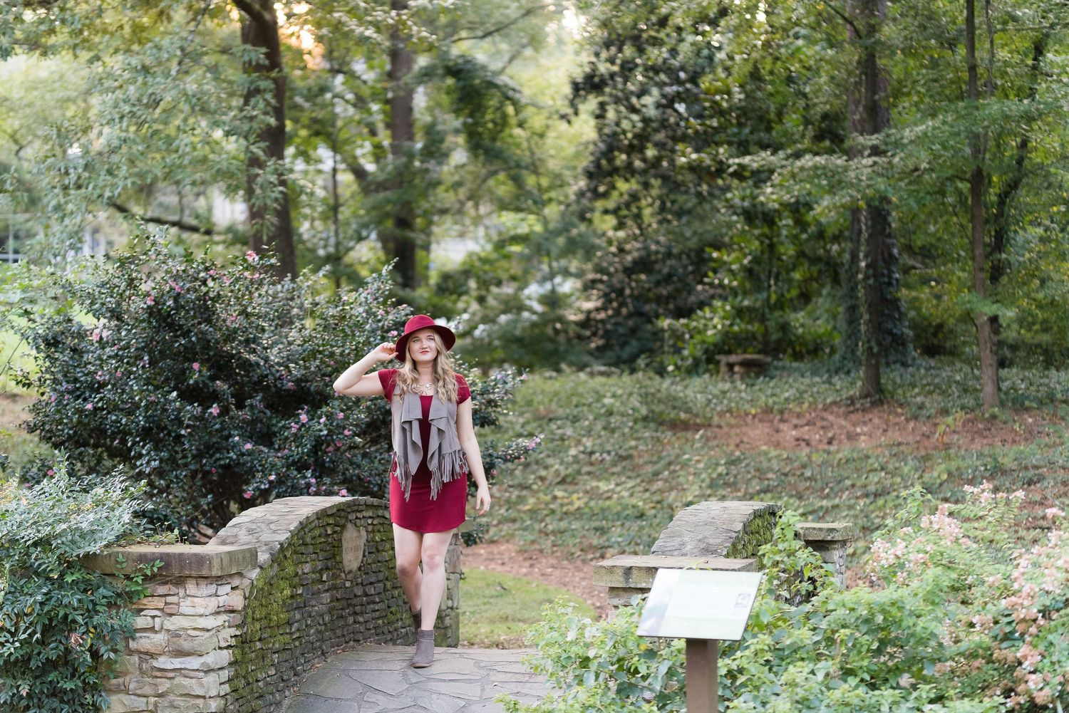 Person in burgundy dress walking along a stone path in a wooded garden setting.