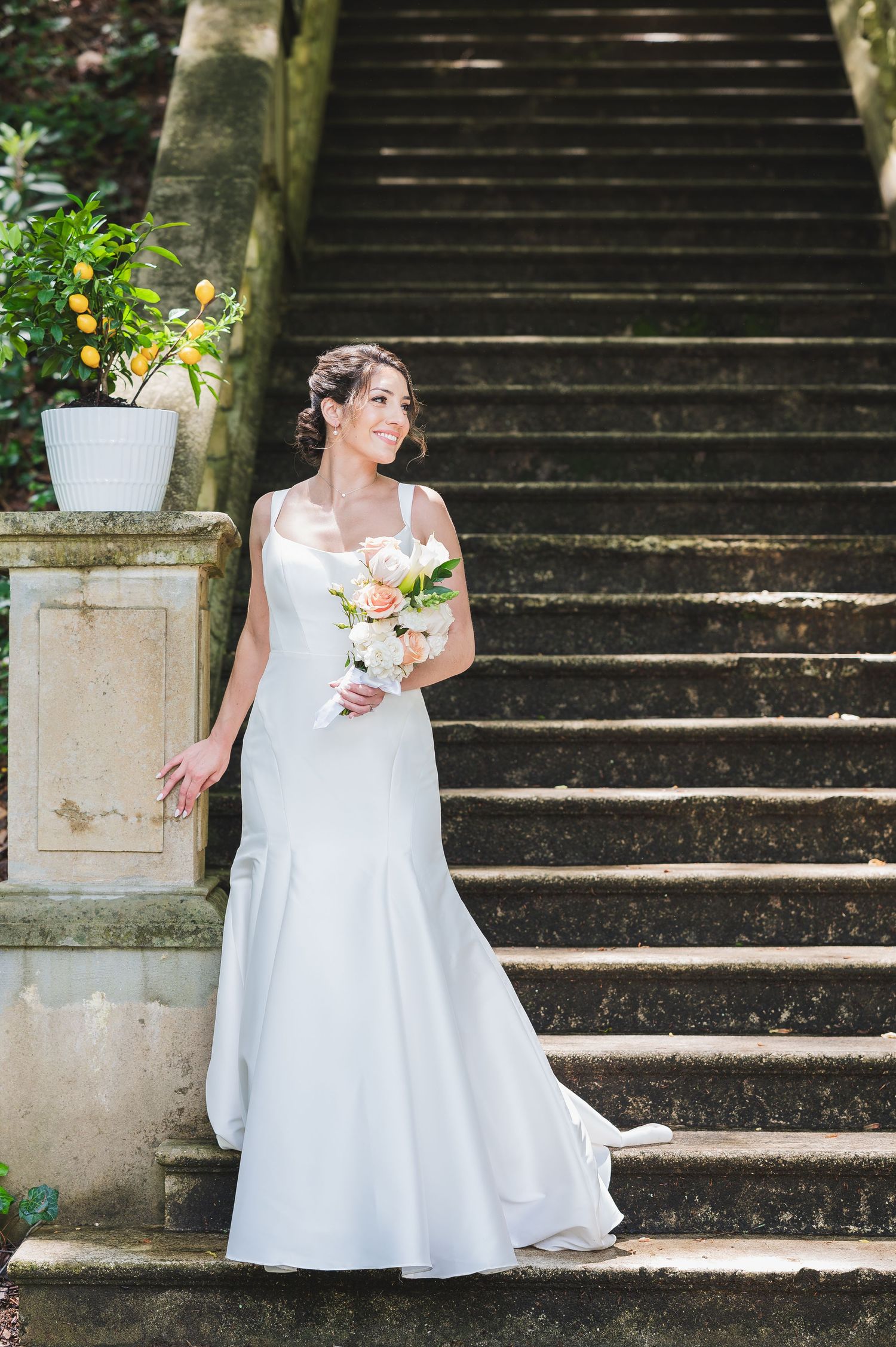 A bride in a flowing white gown poses gracefully on stone steps surrounded by greenery.