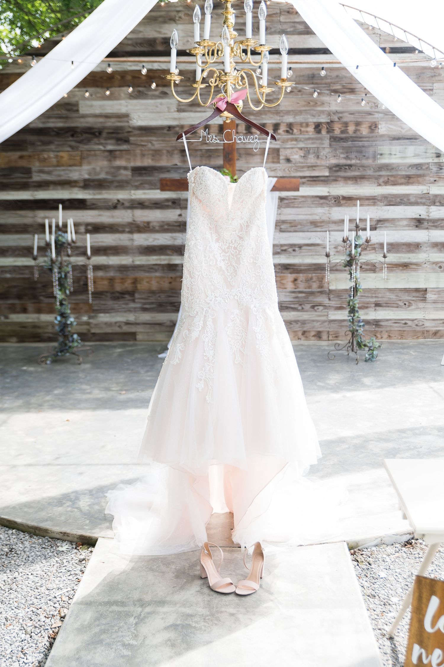 White lace wedding dress hanging on decorative hanger against rustic wooden wall with string lights.