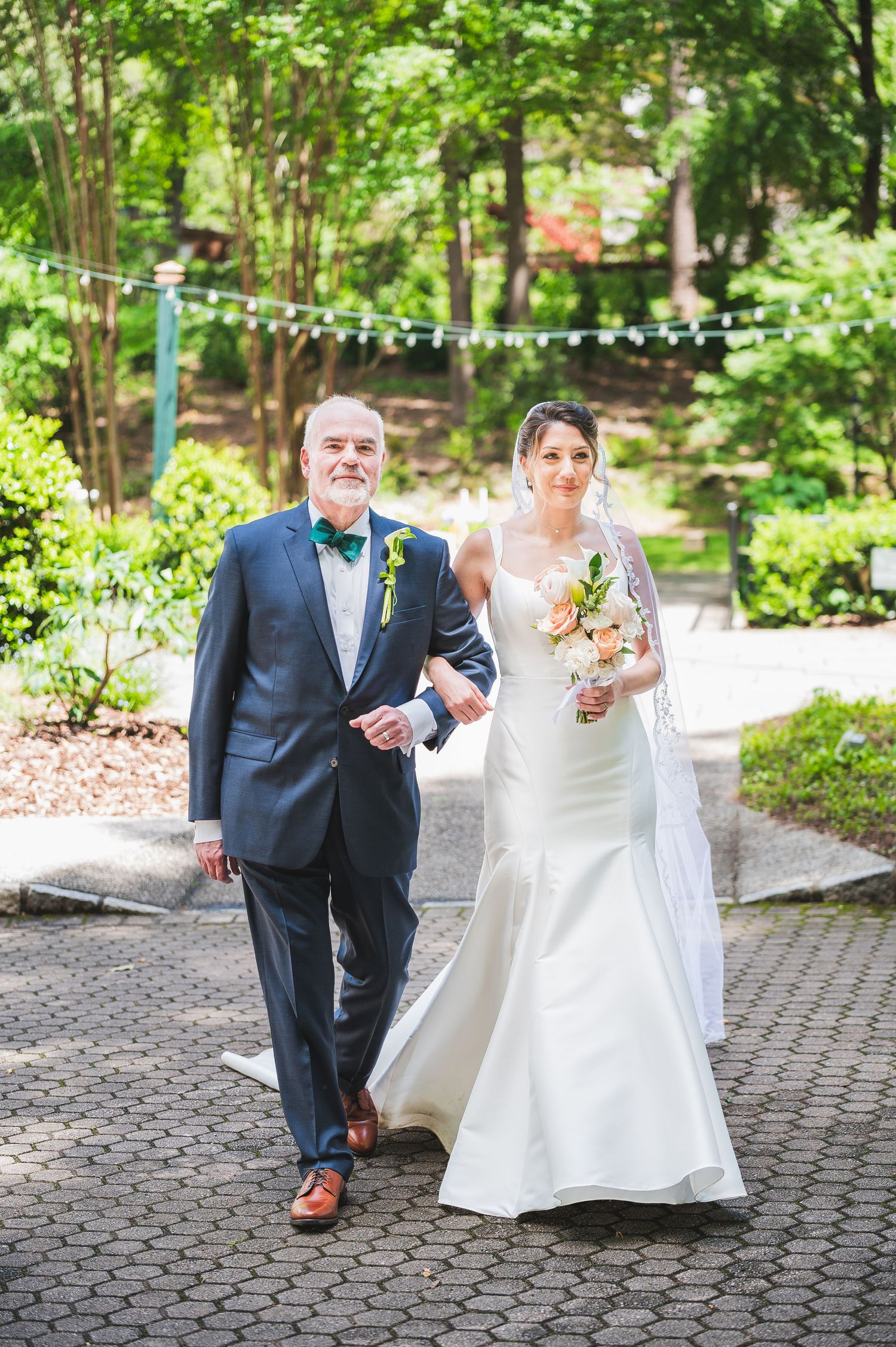 Couple in formal wedding attire walking together on brick pathway through garden setting.