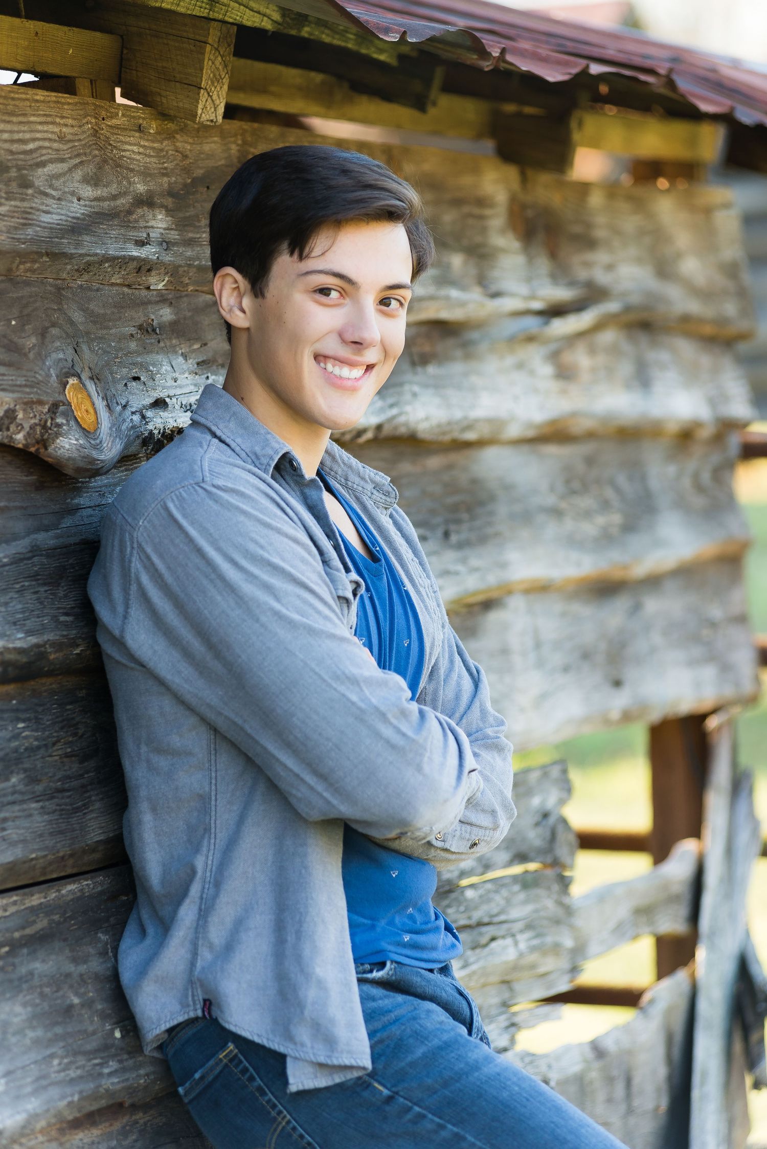 A young man in casual attire leans against a rustic wooden wall while smiling at the camera.