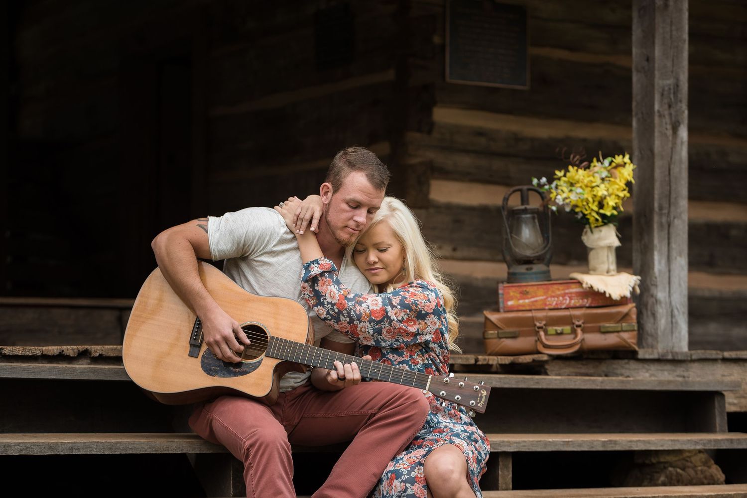 A couple sits close together on rustic cabin steps while playing acoustic guitar with yellow flowers in background.