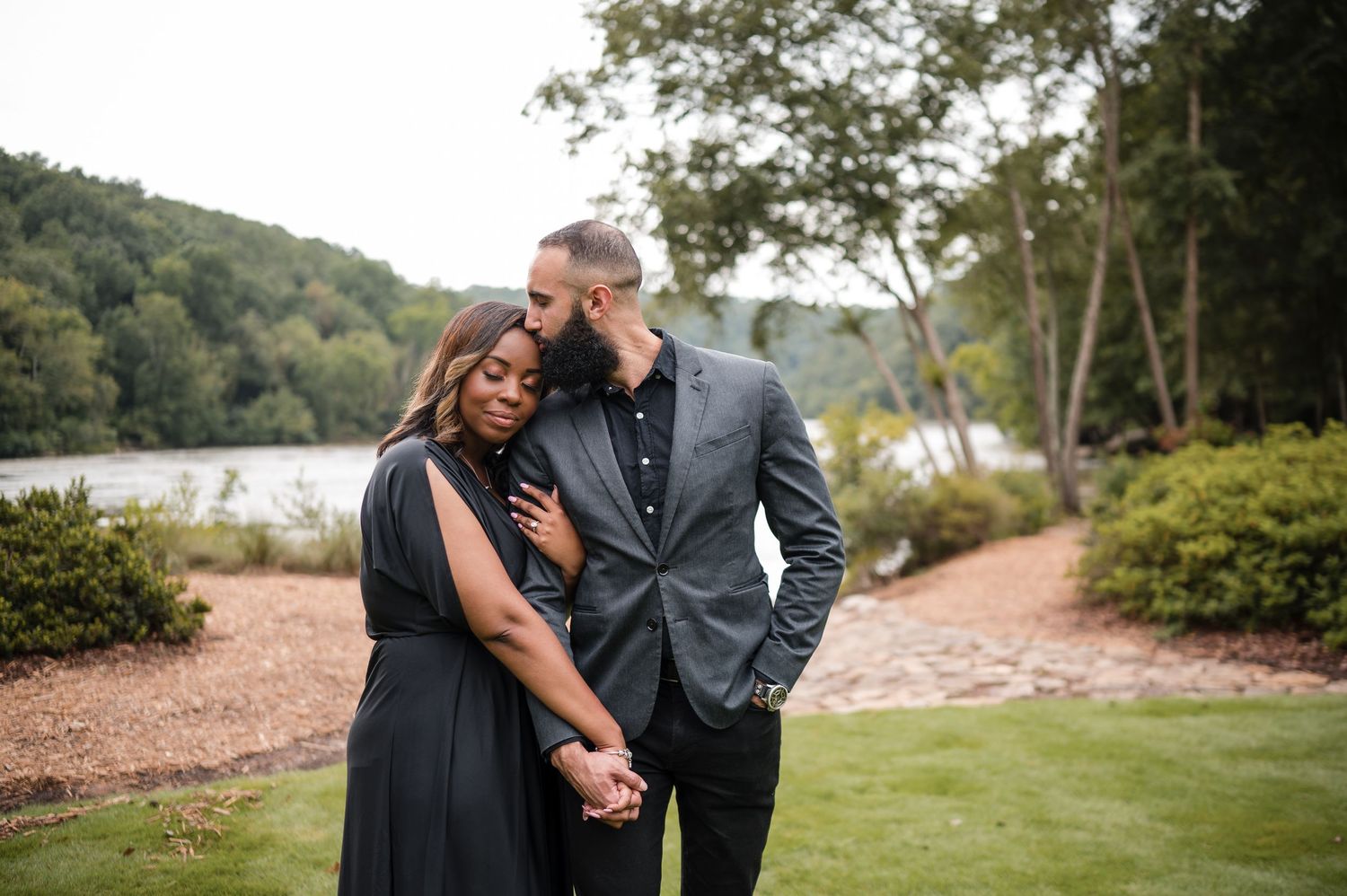 A romantic couple shares an intimate moment by a lakeside path surrounded by lush greenery.