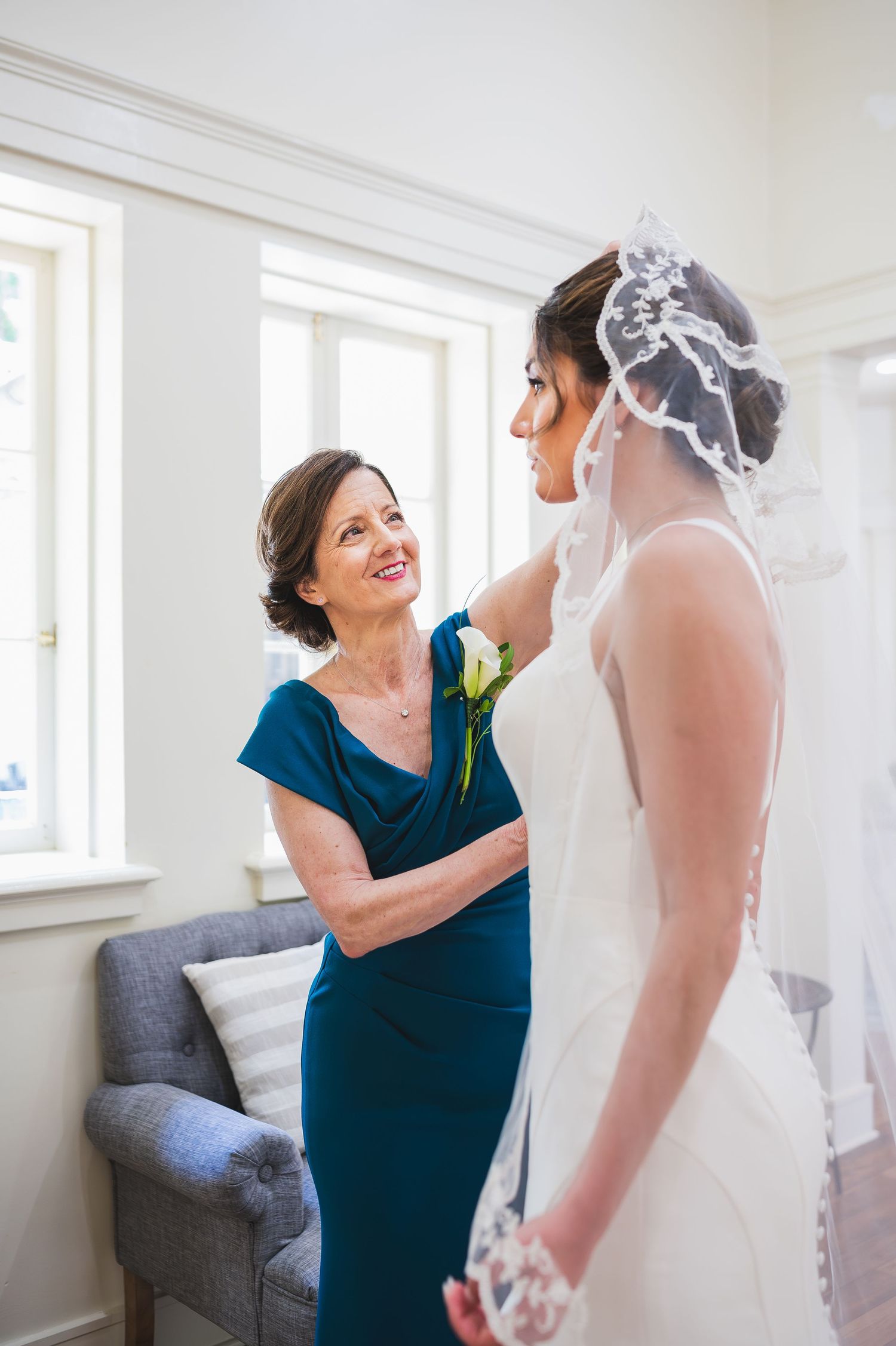 Woman in blue dress helps adjust a wedding gown in a sunlit room.