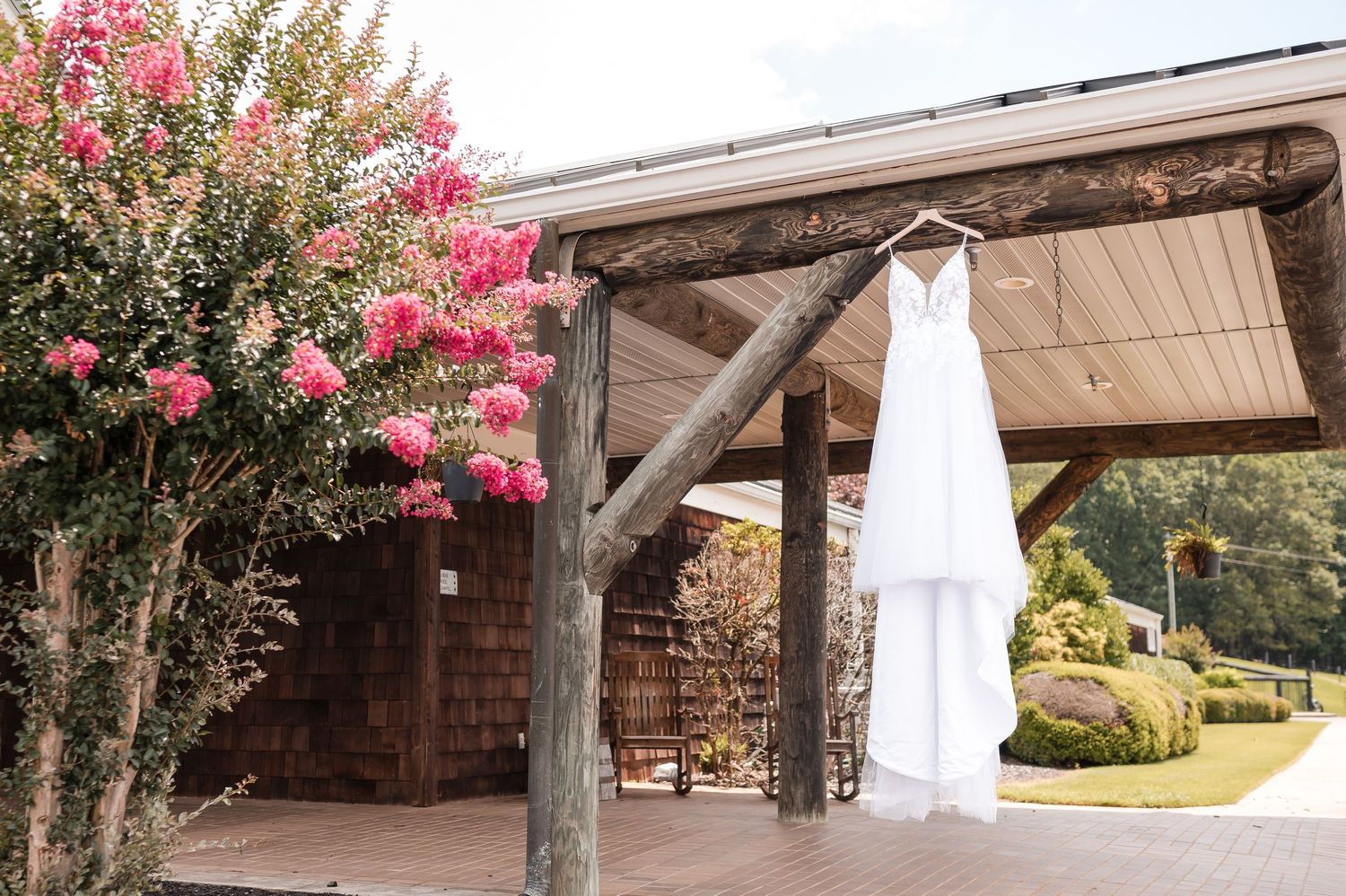 Wedding dress hanging outdoors near blooming pink flowers and rustic architecture.