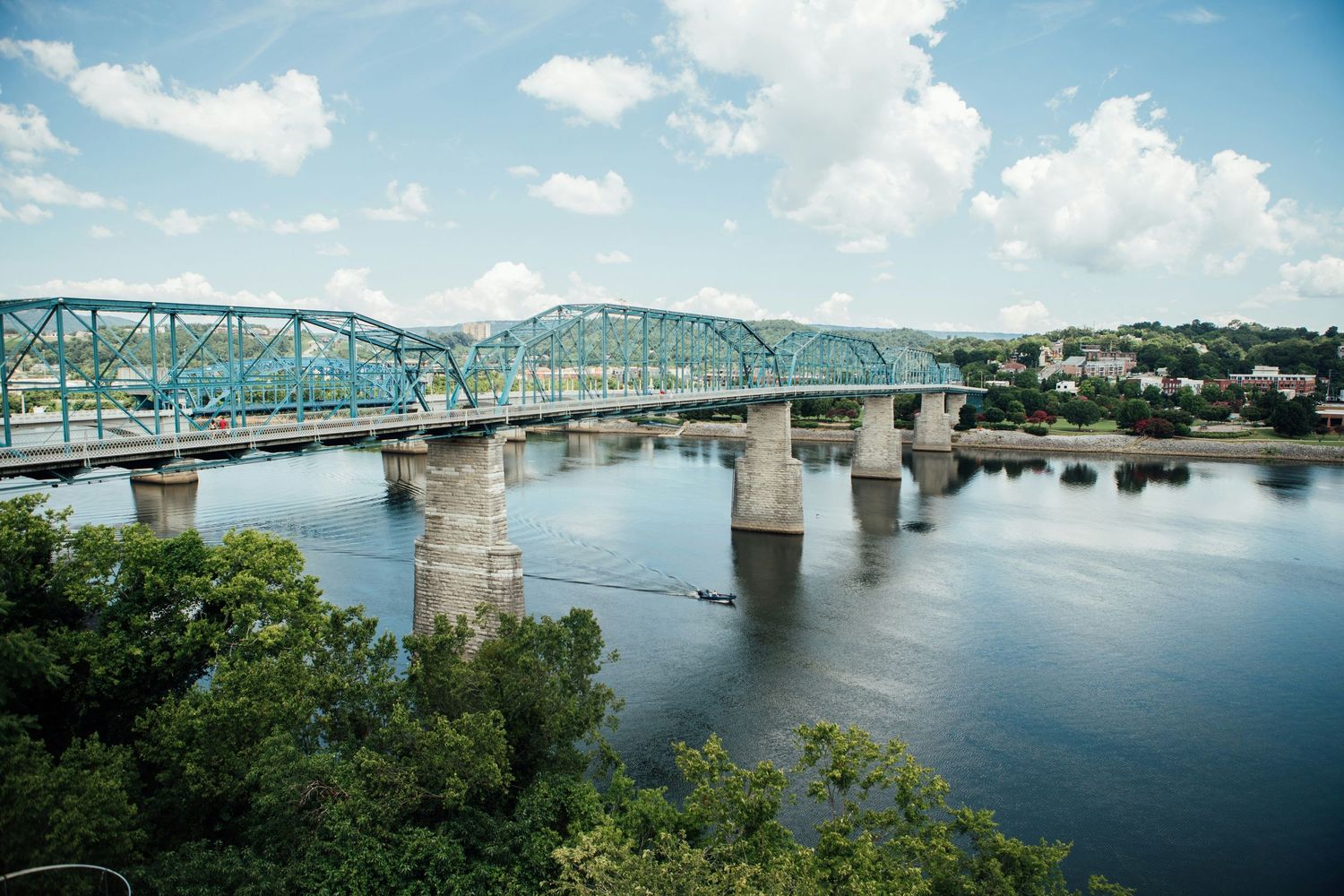 Historic steel truss bridge spanning a calm river under partly cloudy skies, surrounded by lush green trees and hillside town.