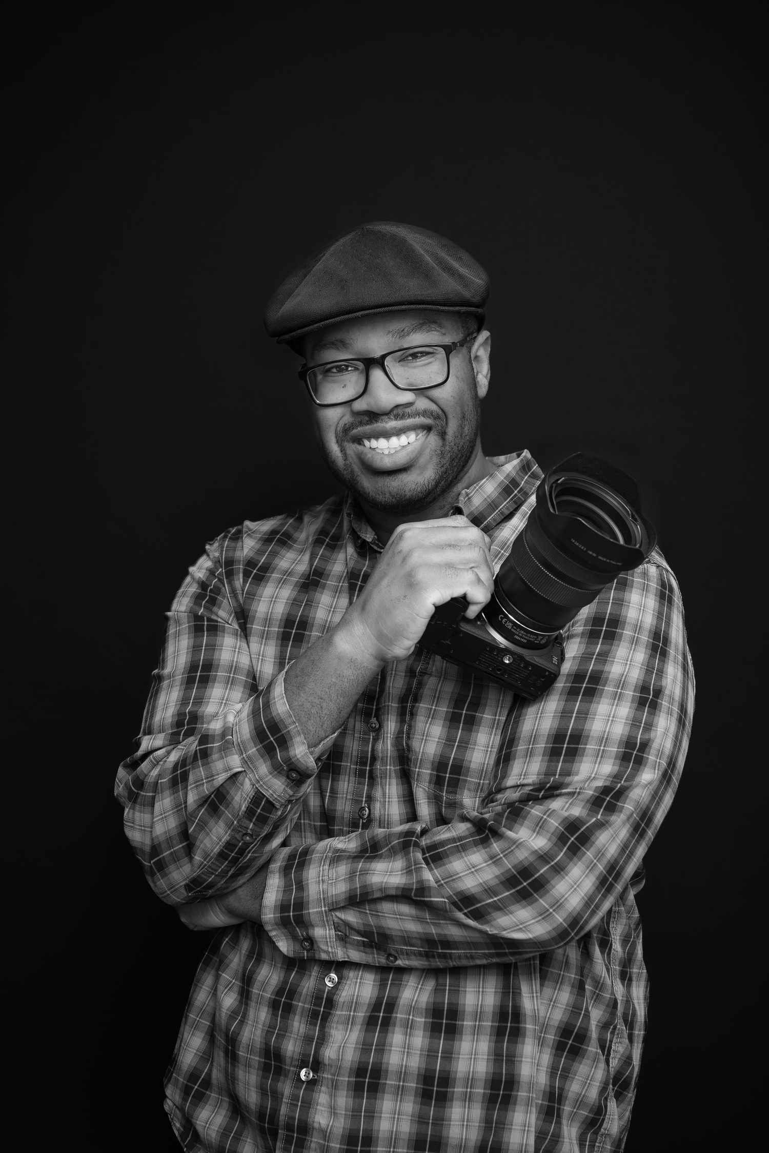 Black and white portrait of photographer wearing plaid shirt, glasses, and cap against dark background.