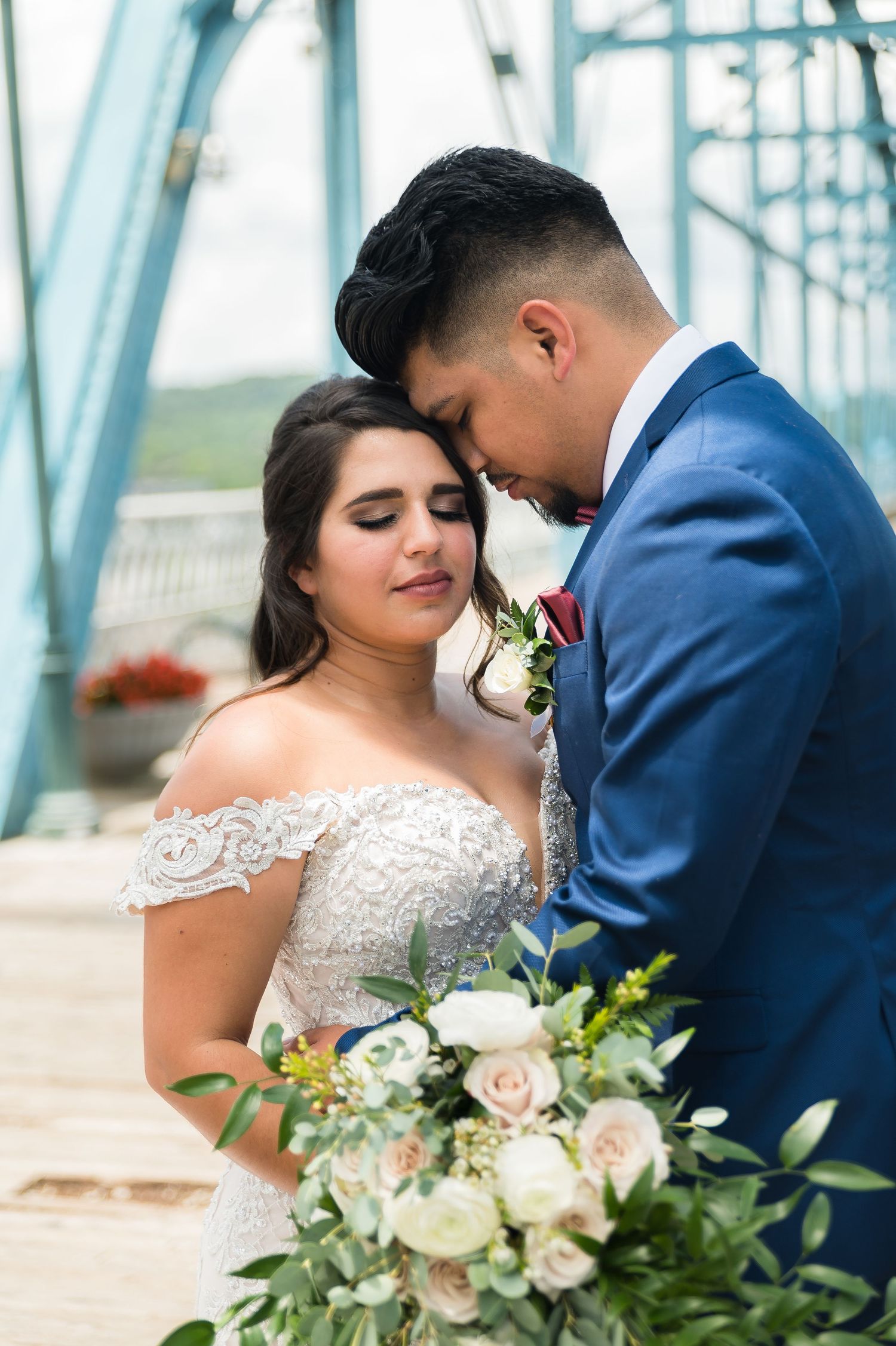 Romantic wedding couple posing by blue bridge with white and blush floral bouquet.