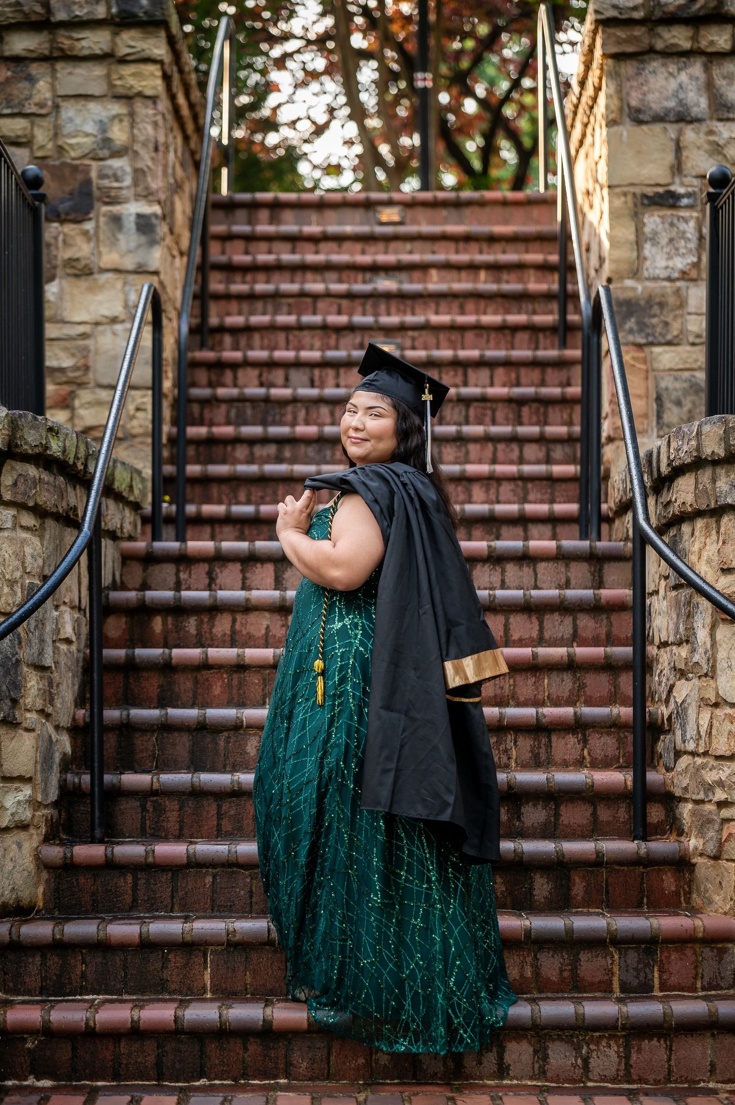 A graduate in a sparkly green dress and black cap and gown poses on stone steps during commencement.