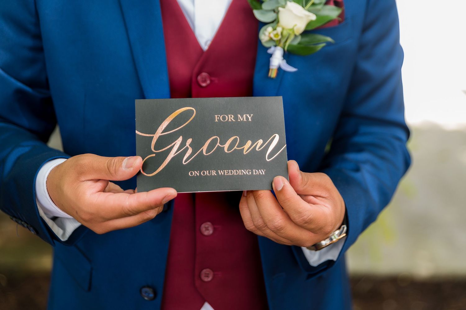 Close up of groom in blue suit holding a small gift box with boutonniere.