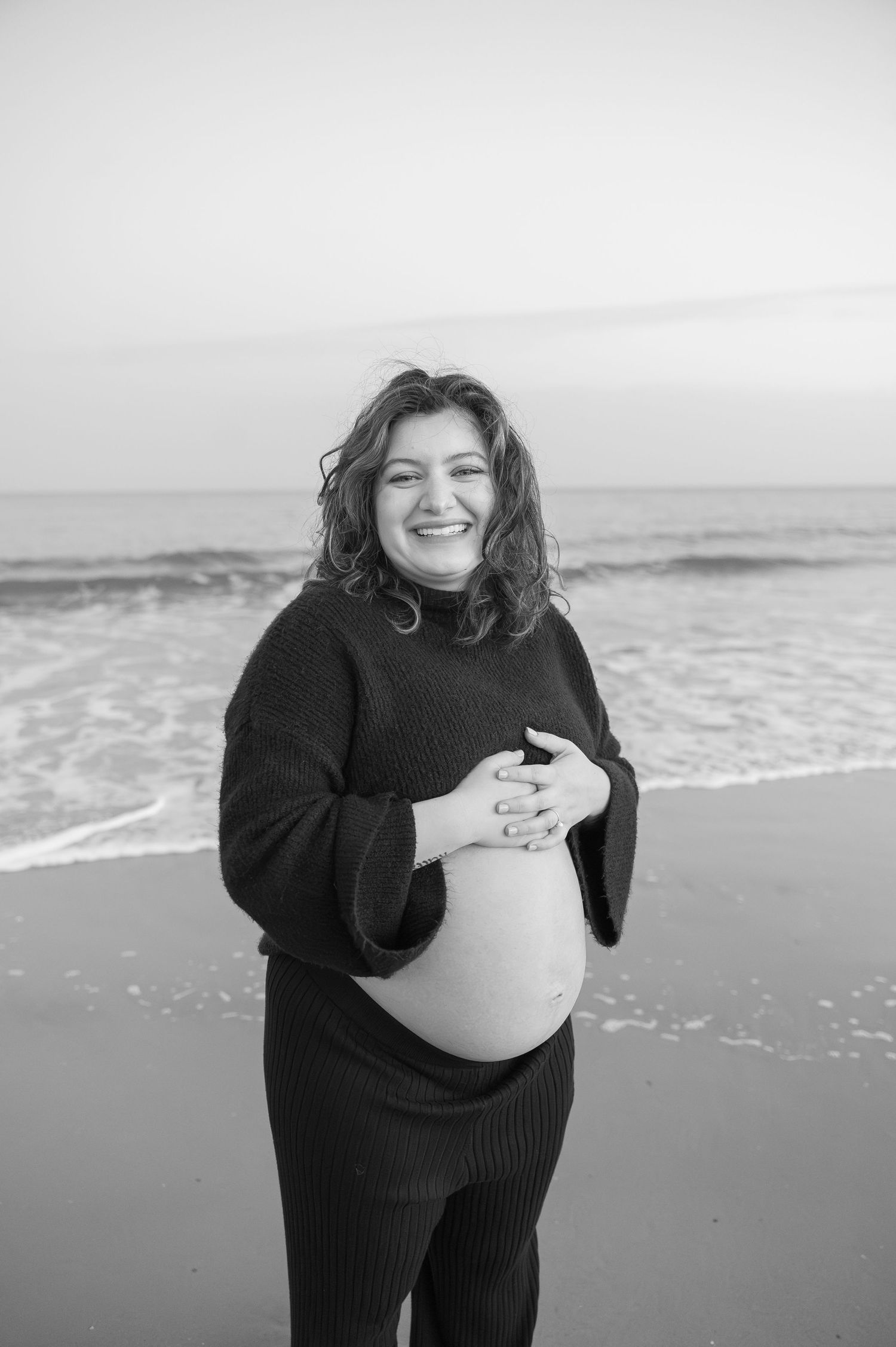 Black and white maternity portraits captured on a beach with ocean waves.