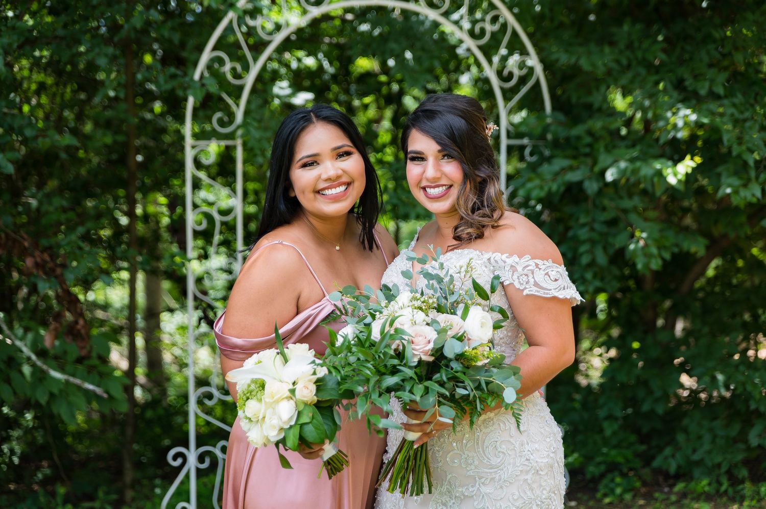 Wedding party members in pastel dresses pose together under a decorative arch outdoors.