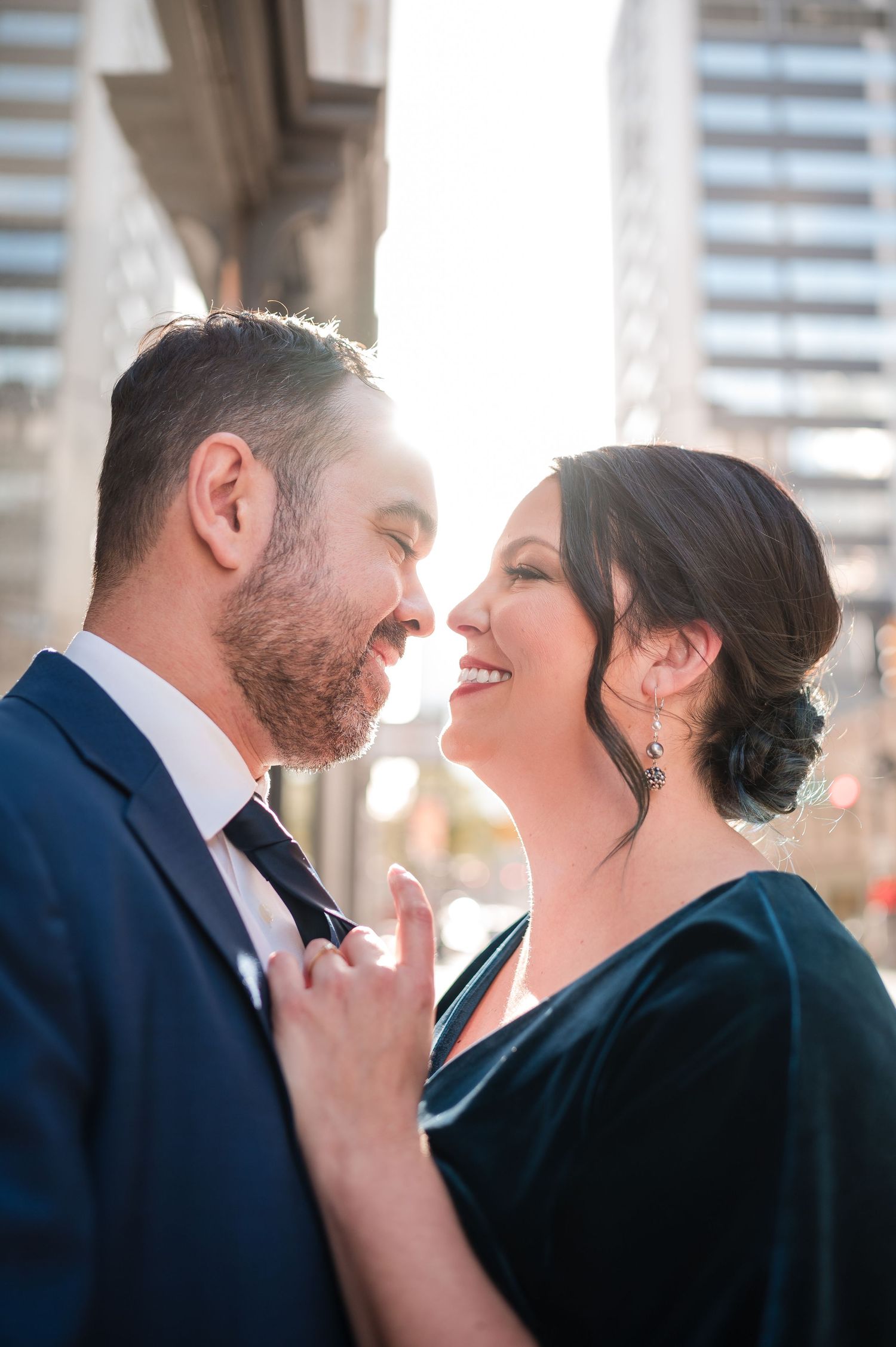 An elegantly dressed couple shares an intimate moment outdoors with city architecture in background.