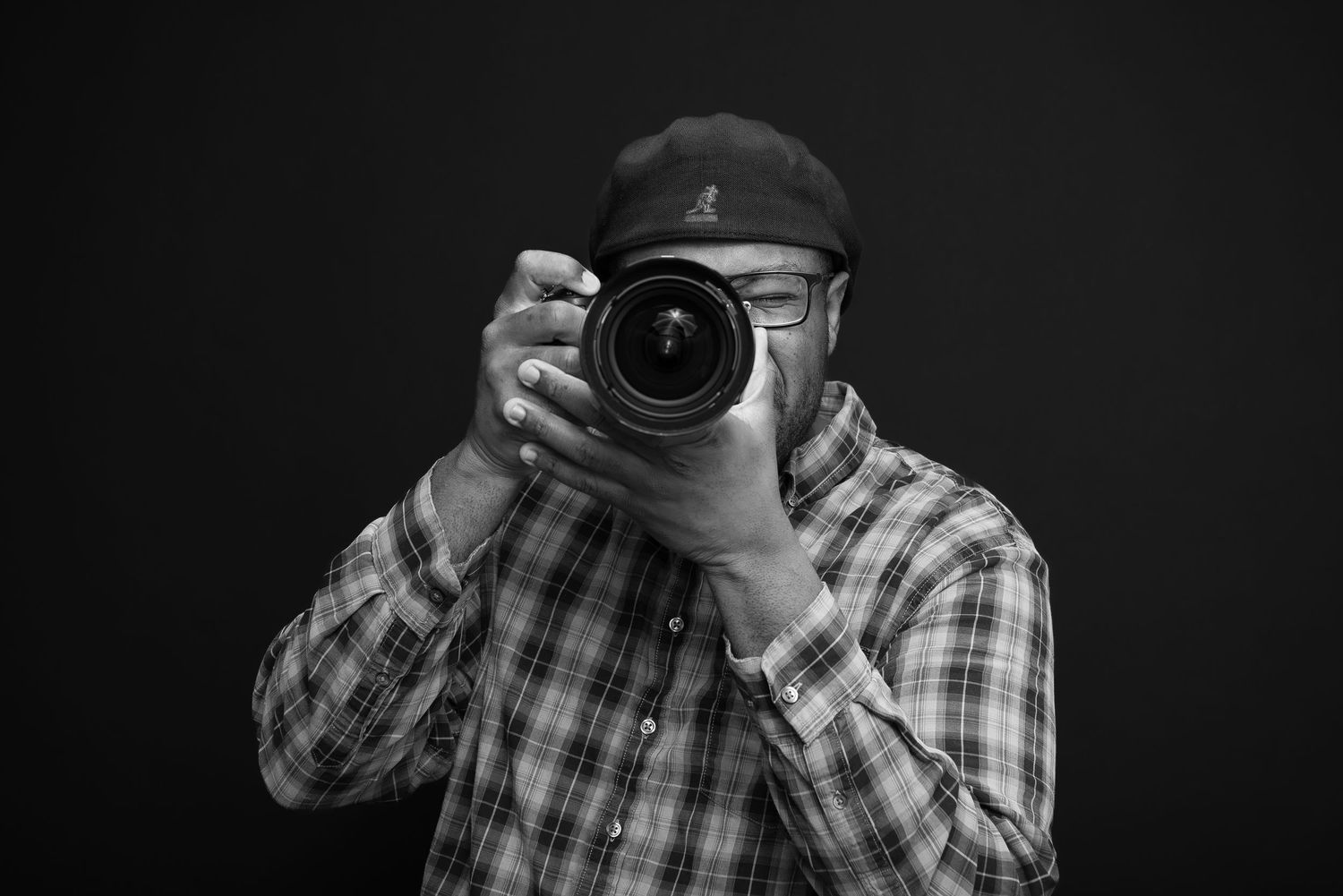 Professional photographer in a plaid shirt capturing photos with a DSLR camera against dark background.