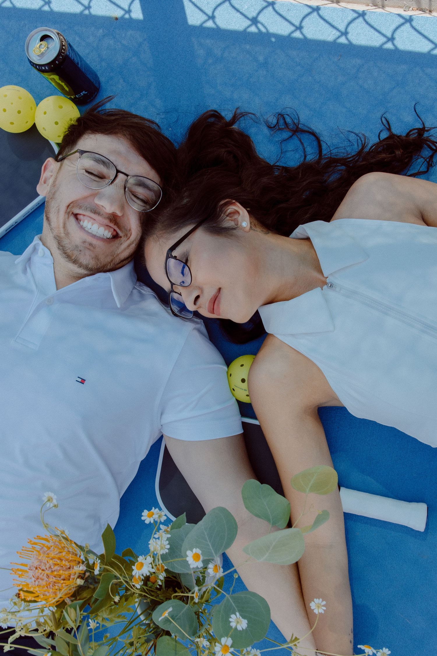 A couple lies on a  blue tennis court surrounded by delicate white and yellow flowers in an intimate portrait.