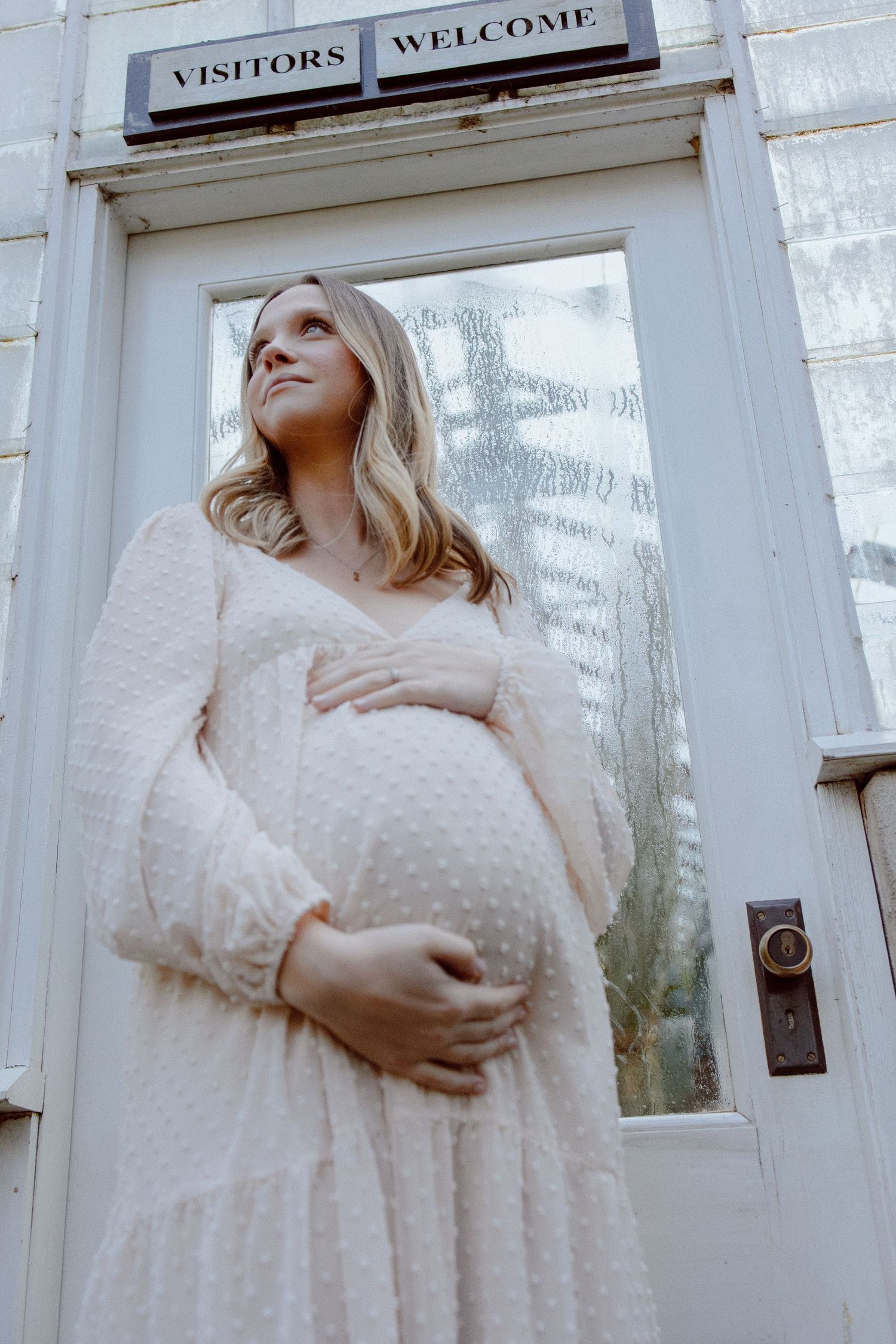 An expecting mother in a pale knit dress posing against a white greenhouse building with a glass window door.