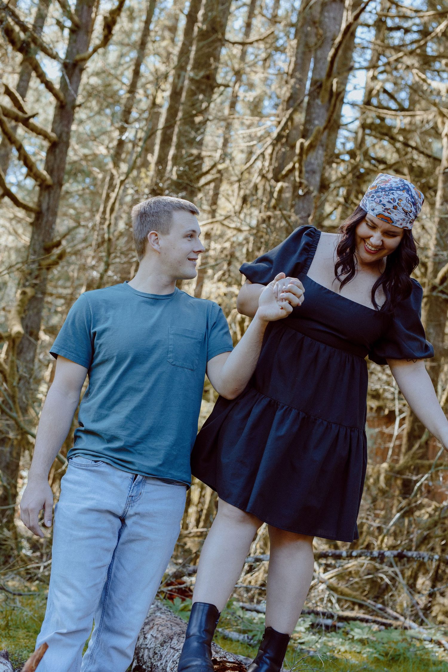 A couple wearing blue and black outfits holds hands while walking through a sunlit wooded area.