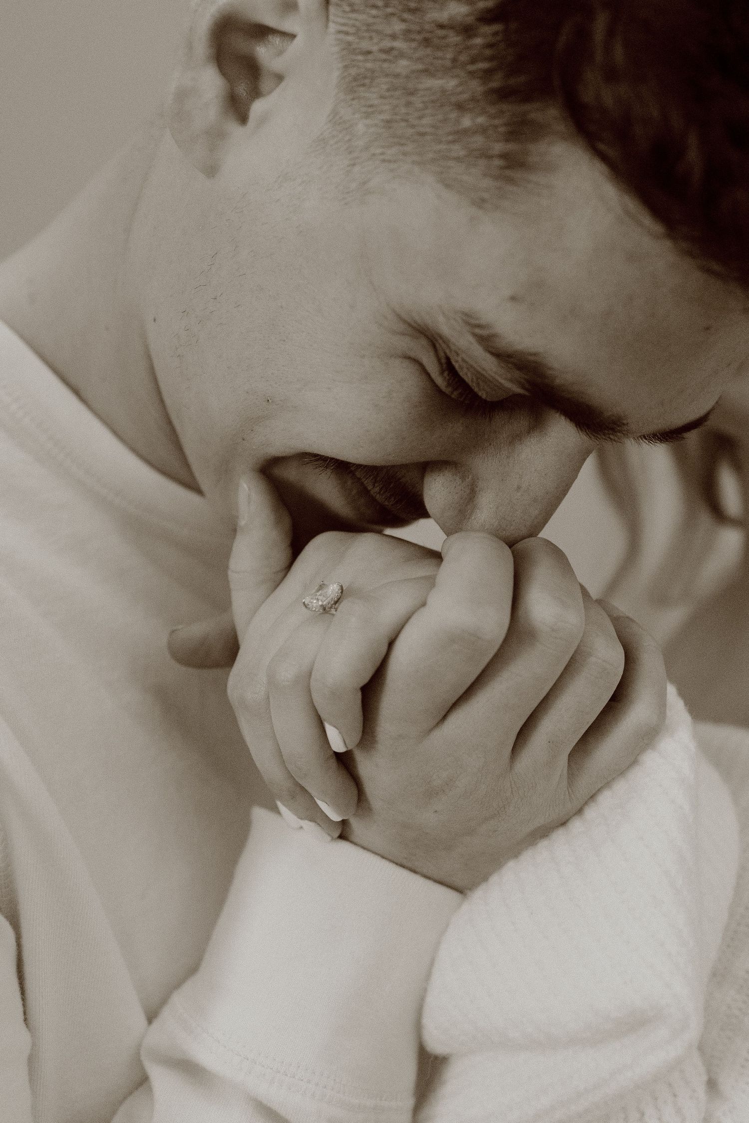 Close-up artistic black and white photograph showing hands clasped near face, with fiancée kissing the hand with wedding ring atop it.