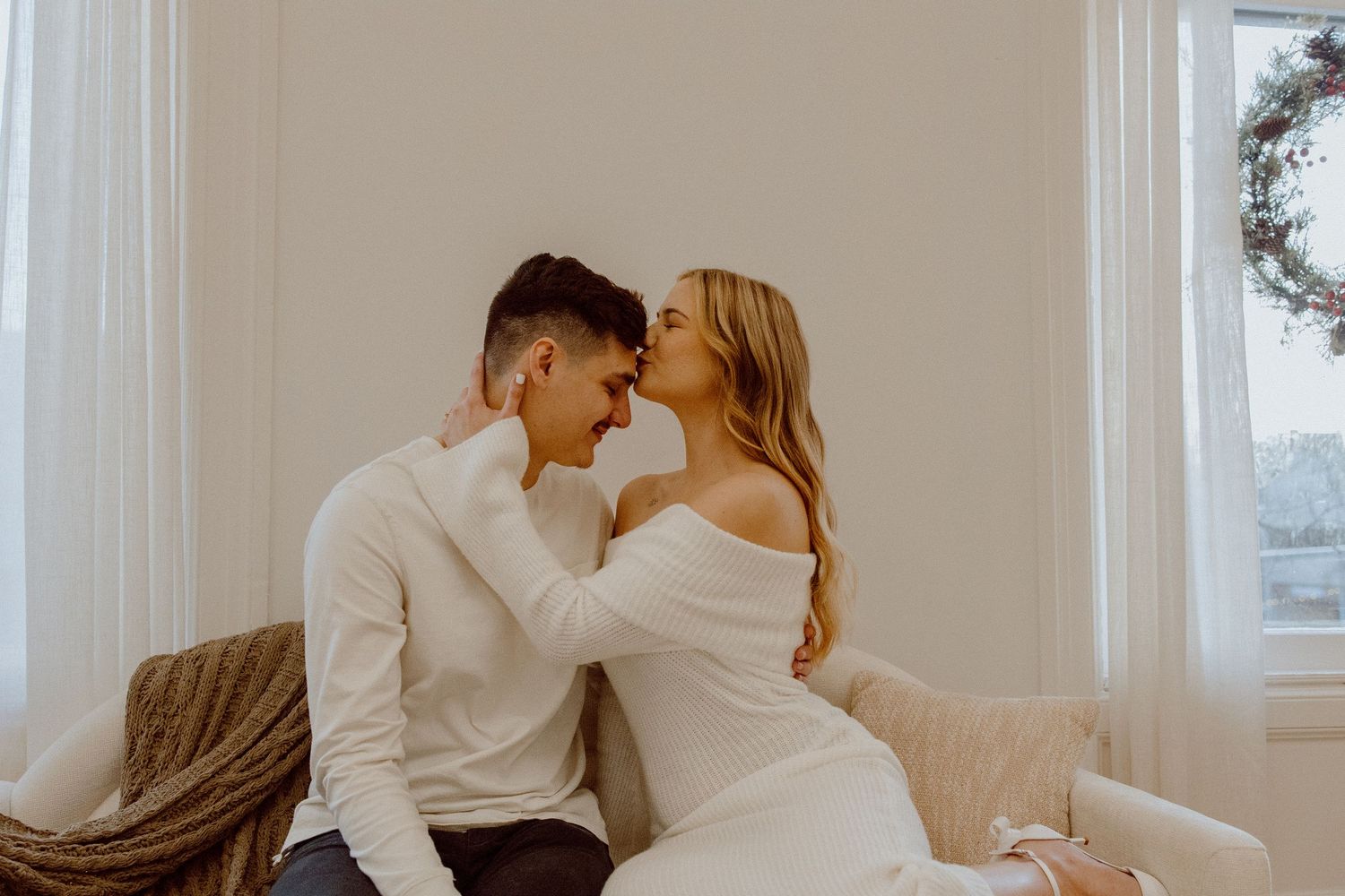 Romantic couple in stylish wedding attire sharing intimate moments on a couch in a bright, sunlit room.
