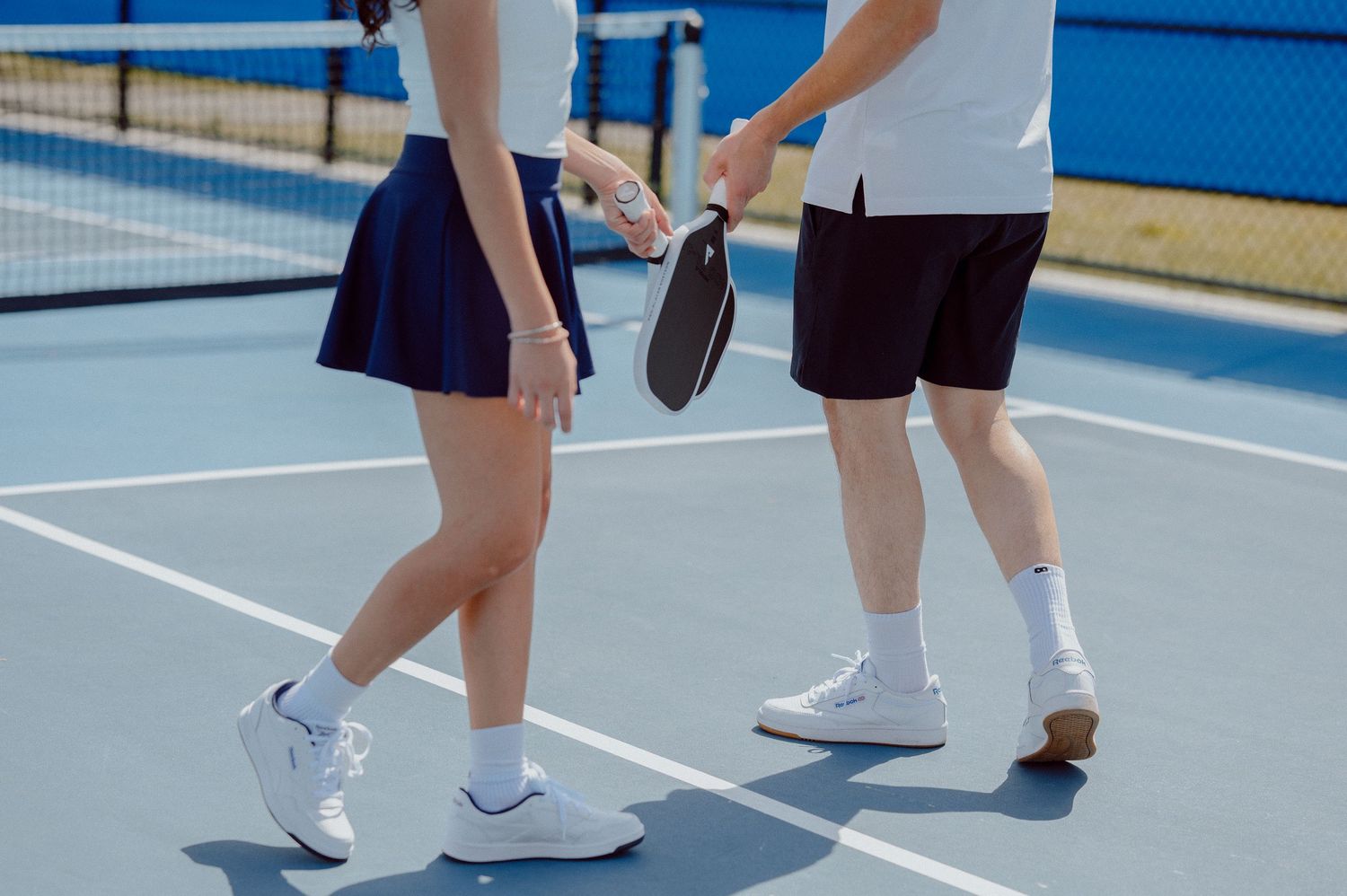 Close-up view of players' feet and legs on a blue tennis court during a recreational pickle ball match.