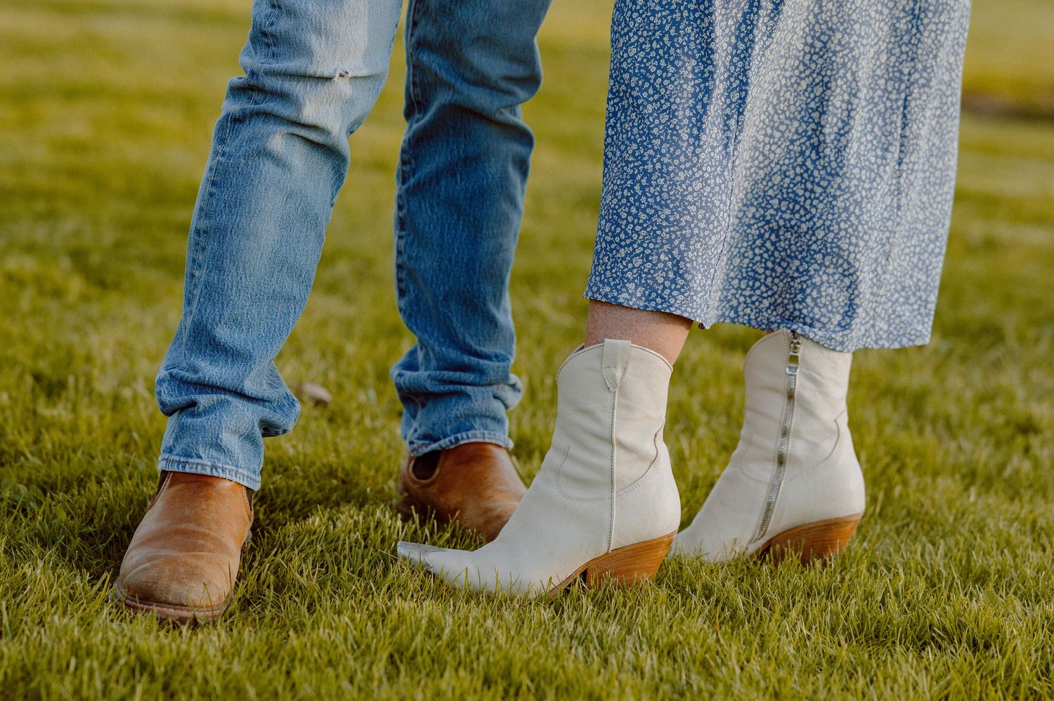 Row of feet wearing brown and white western boots on green grass in a casual outdoor setting.