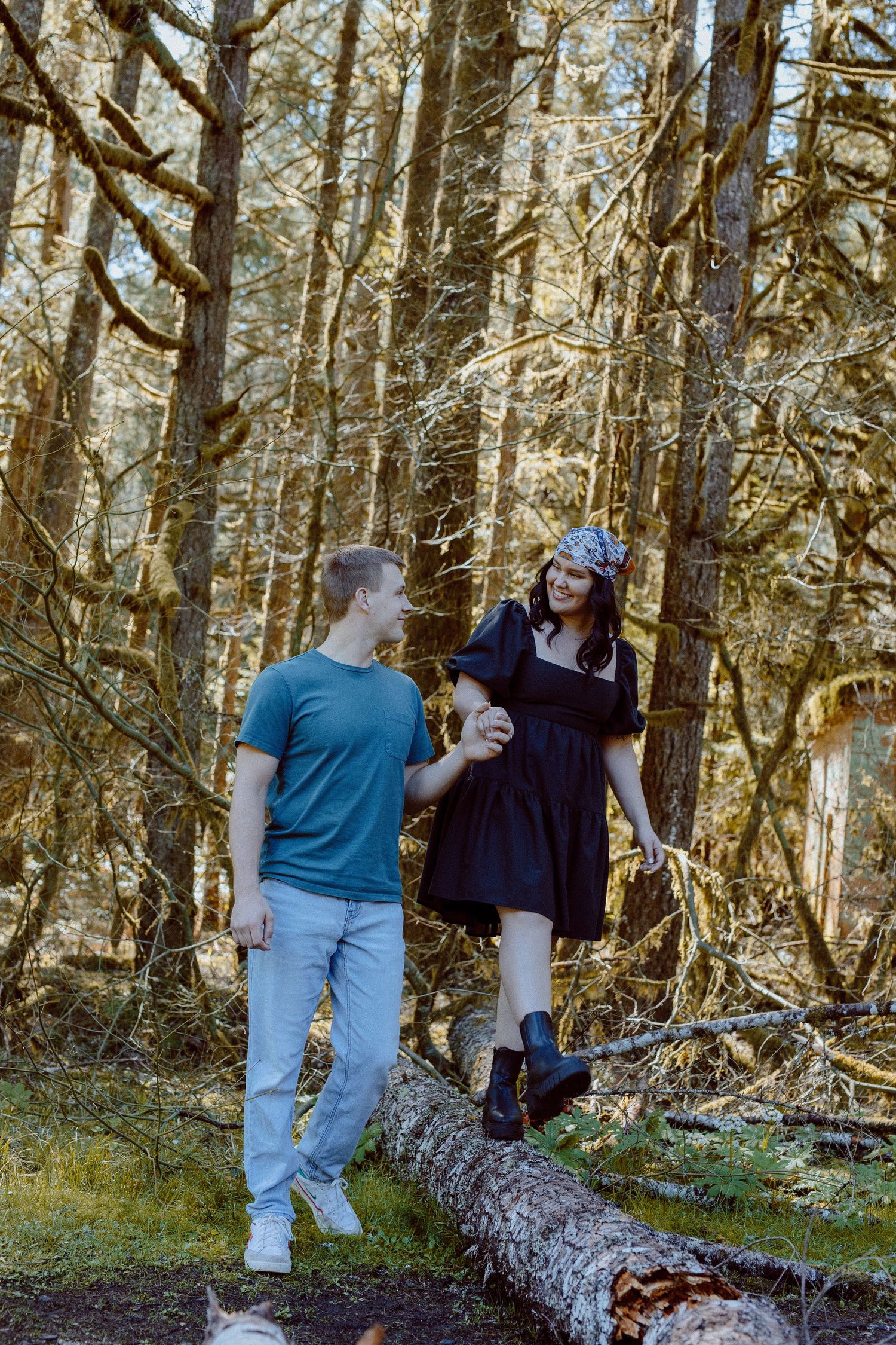 A couple shares a moment together while exploring a mossy forest with fallen logs and sunlit trees in the background.