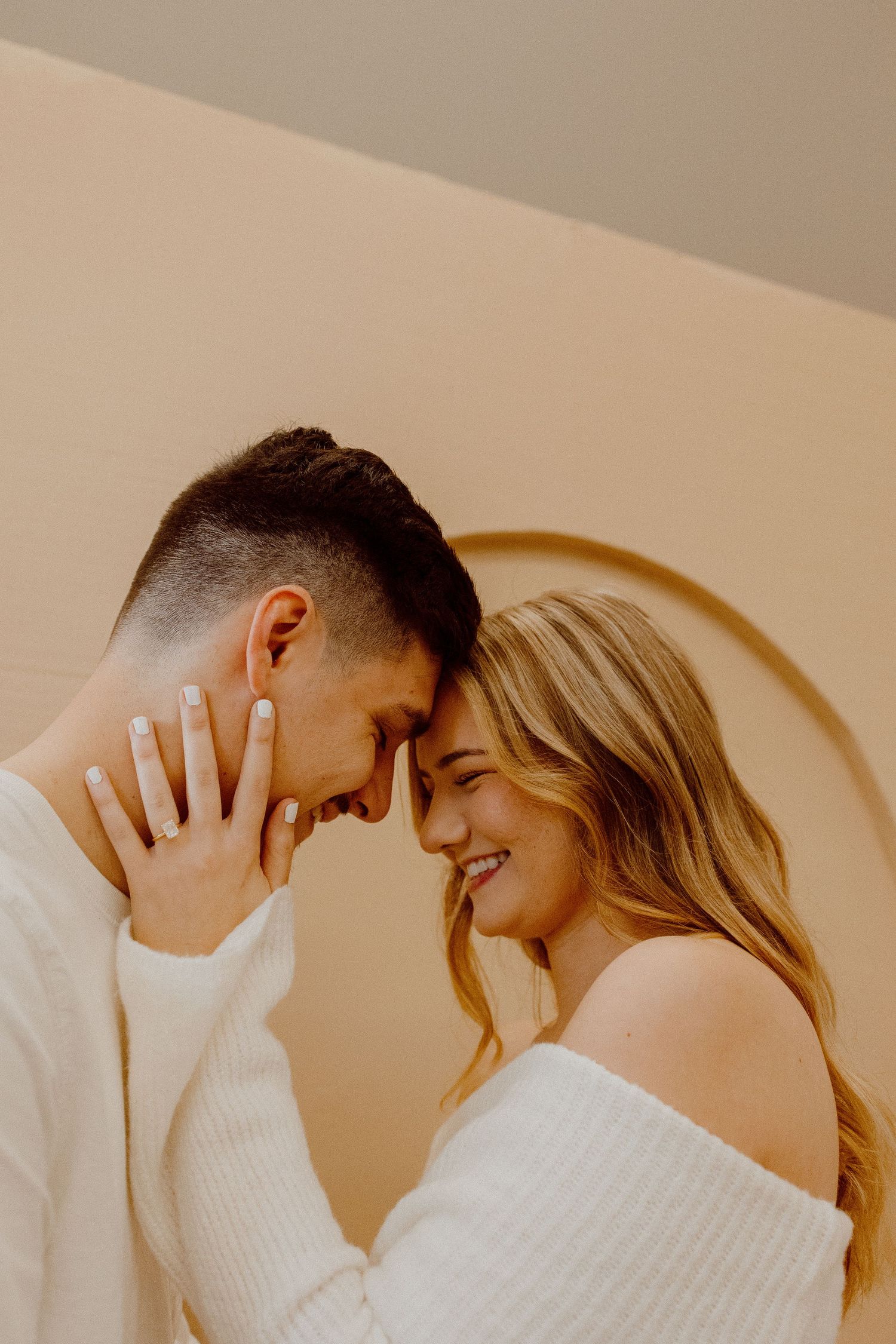 A couple in white sweaters share an intimate candid moment together against a neutral wall background.